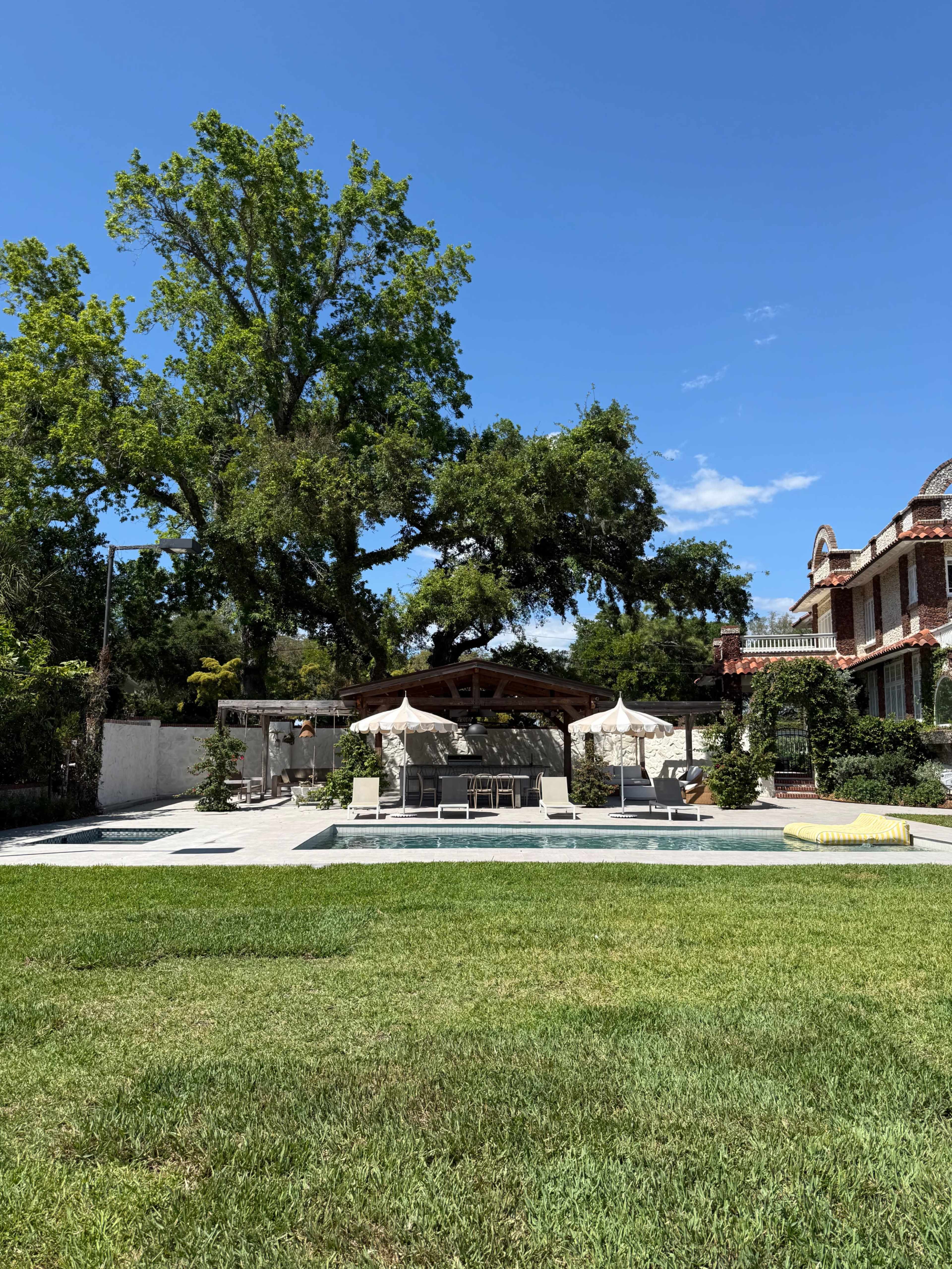 The image shows a spacious backyard with a swimming pool, outdoor seating under large umbrellas, and a well-maintained lawn, set against a clear blue sky and a house in the background.