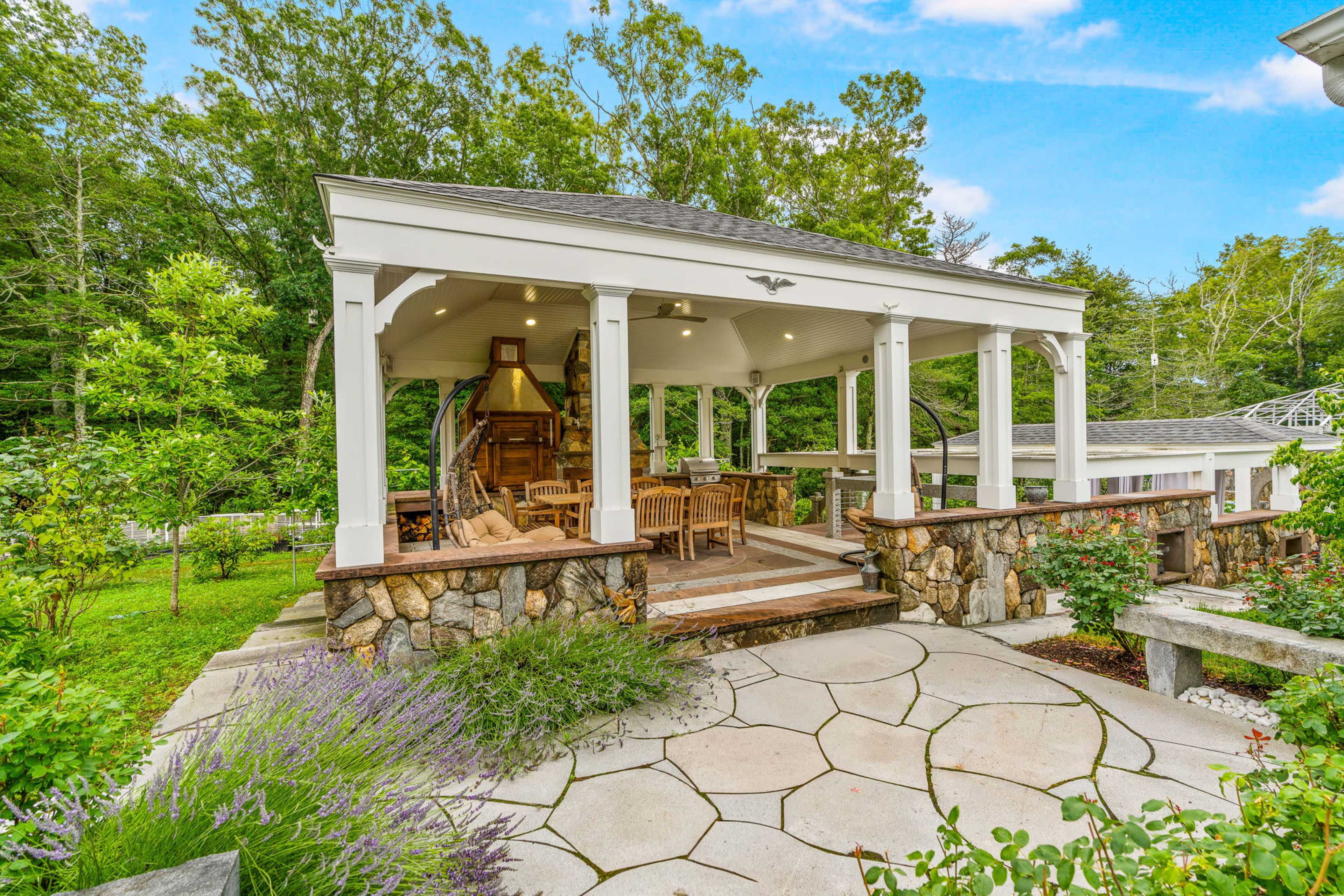 The image shows a wooden gazebo with a stone foundation, surrounded by greenery and a patterned stone path.