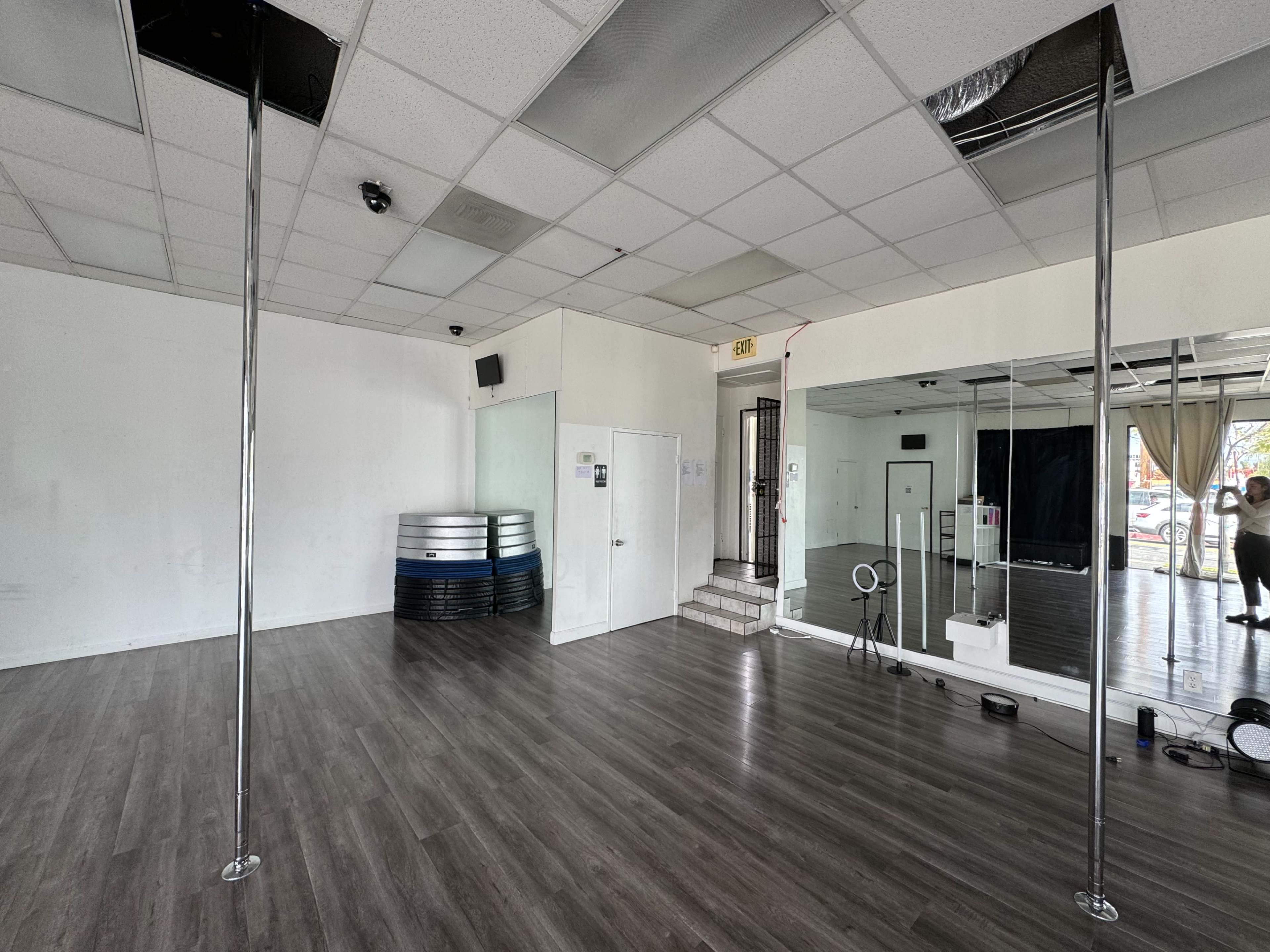 A spacious, empty studio with two pole dance poles, a mirrored wall, and a staircase leading to an exit.
