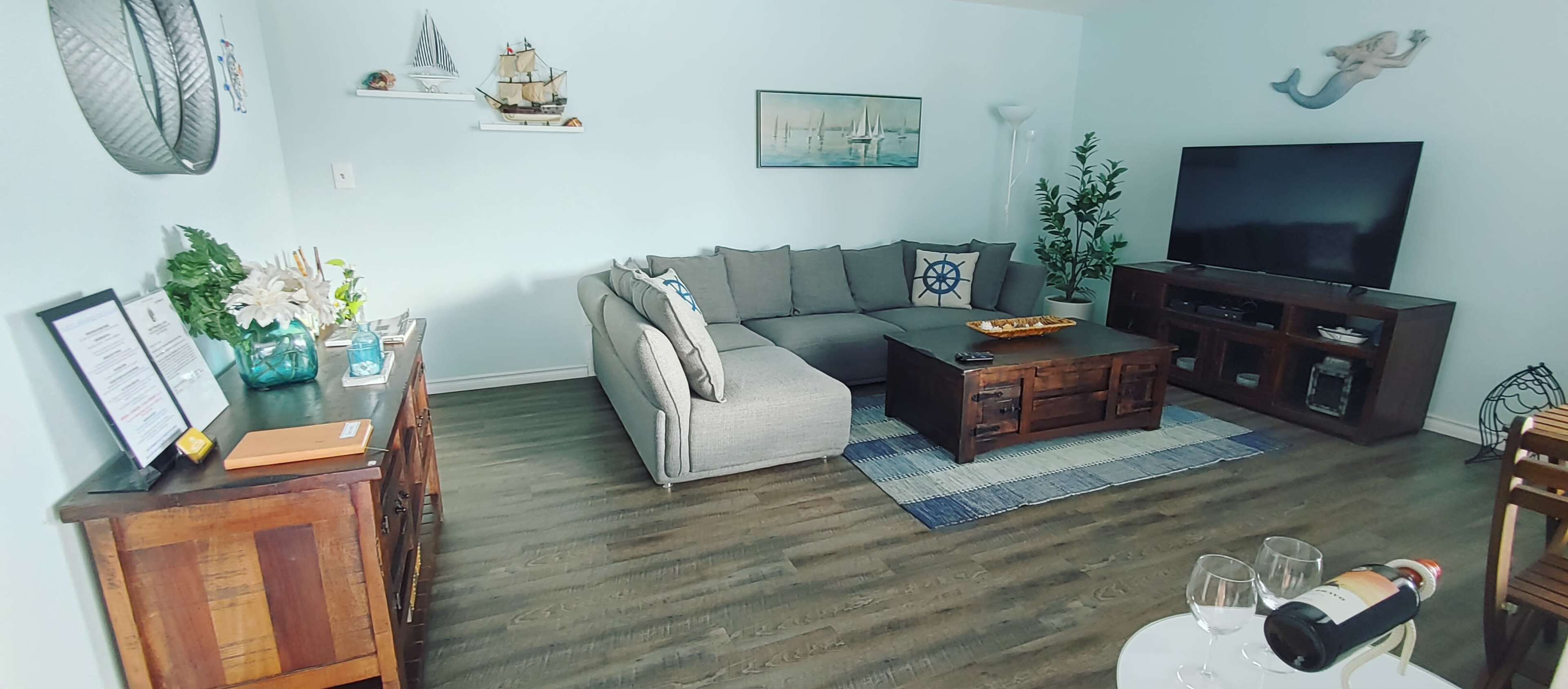 The image shows a stylish living room with a sectional sofa, a wooden coffee table, a television, and nautical-themed decor.