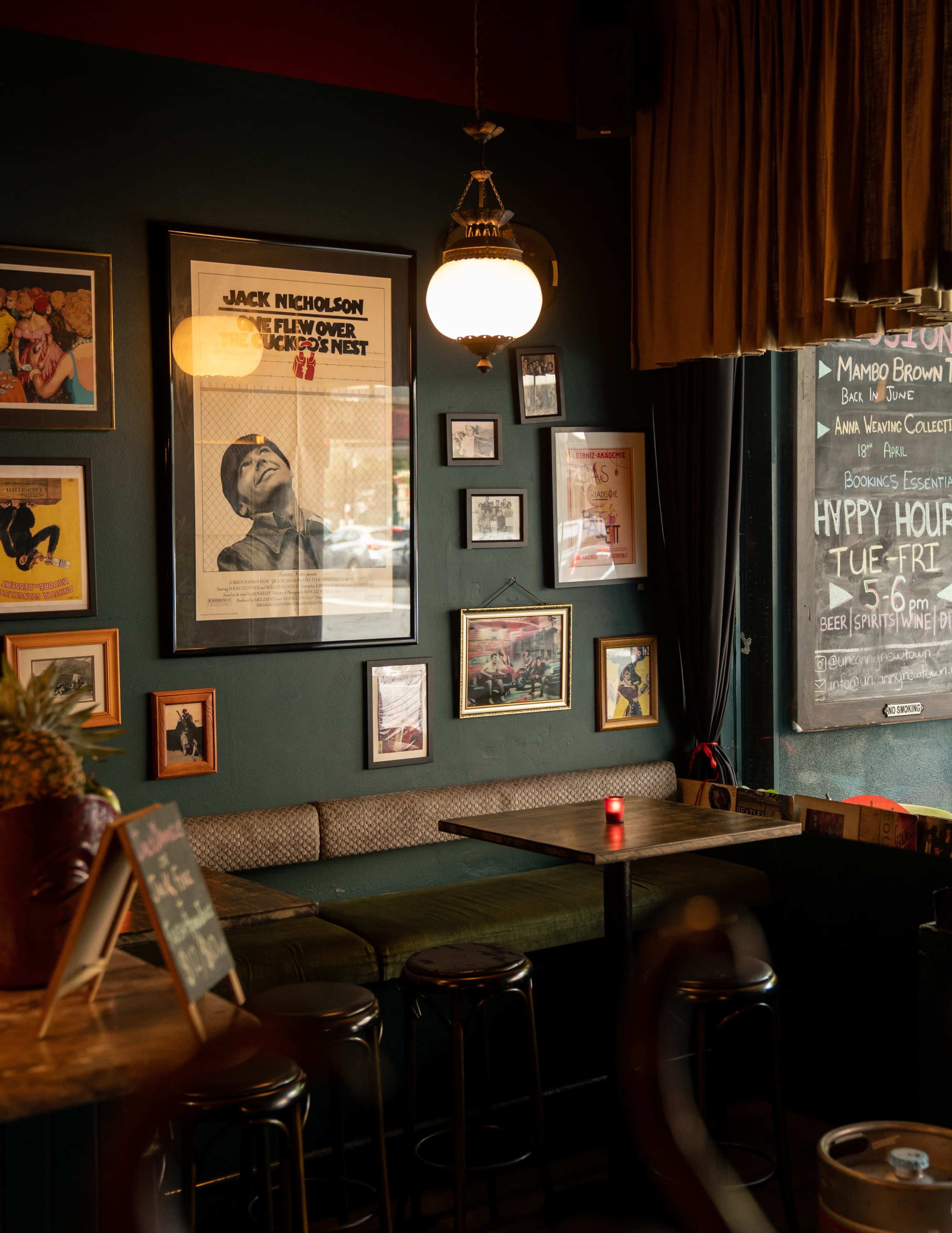 The image shows a cozy bar interior featuring dark green walls adorned with framed movie posters, a vintage lamp hanging from the ceiling, and a wooden table with high stools.