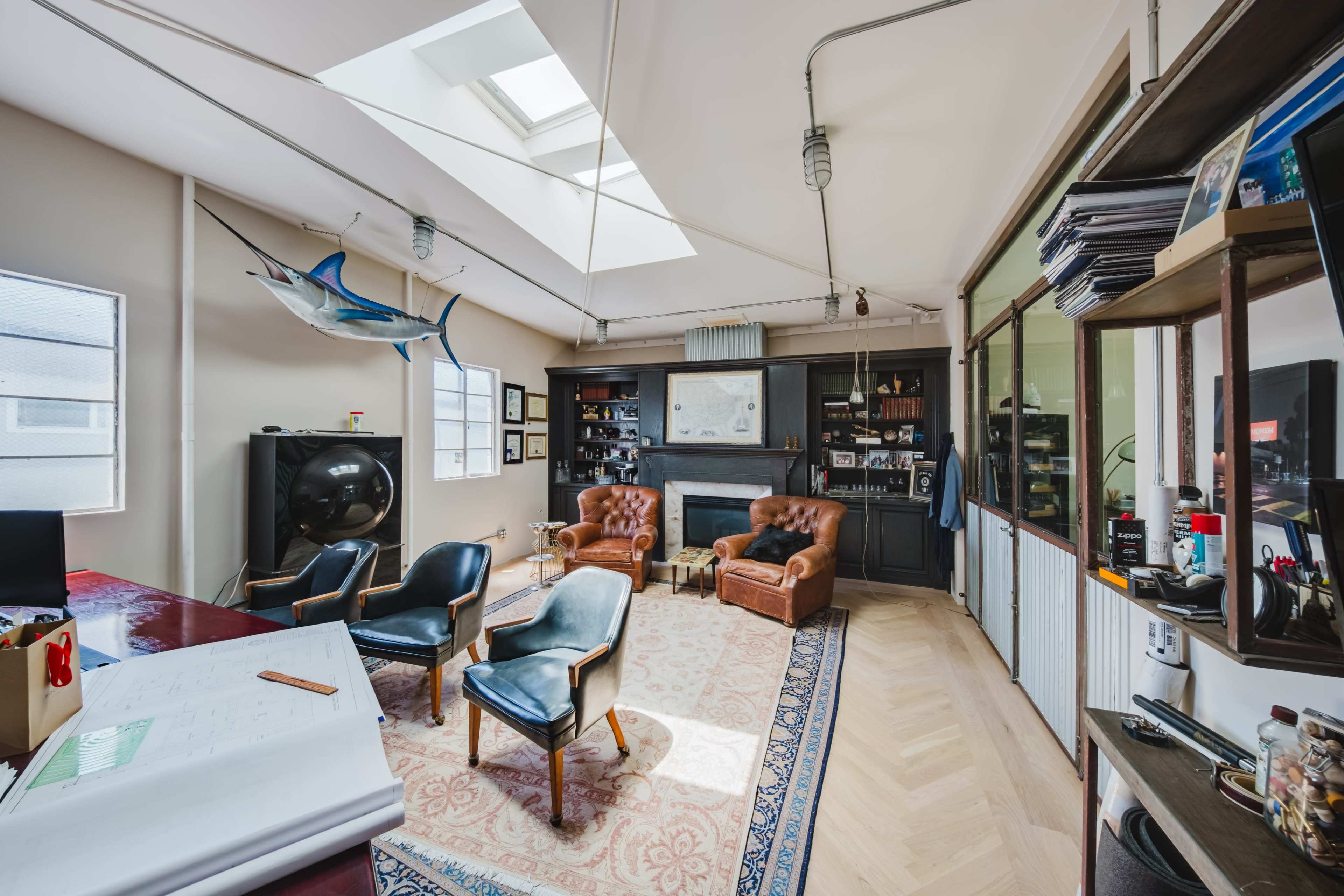 The image shows a modern office space featuring two brown leather armchairs, a black television, and a large fish mounted on the wall, with a central skylight allowing natural light to enter.
