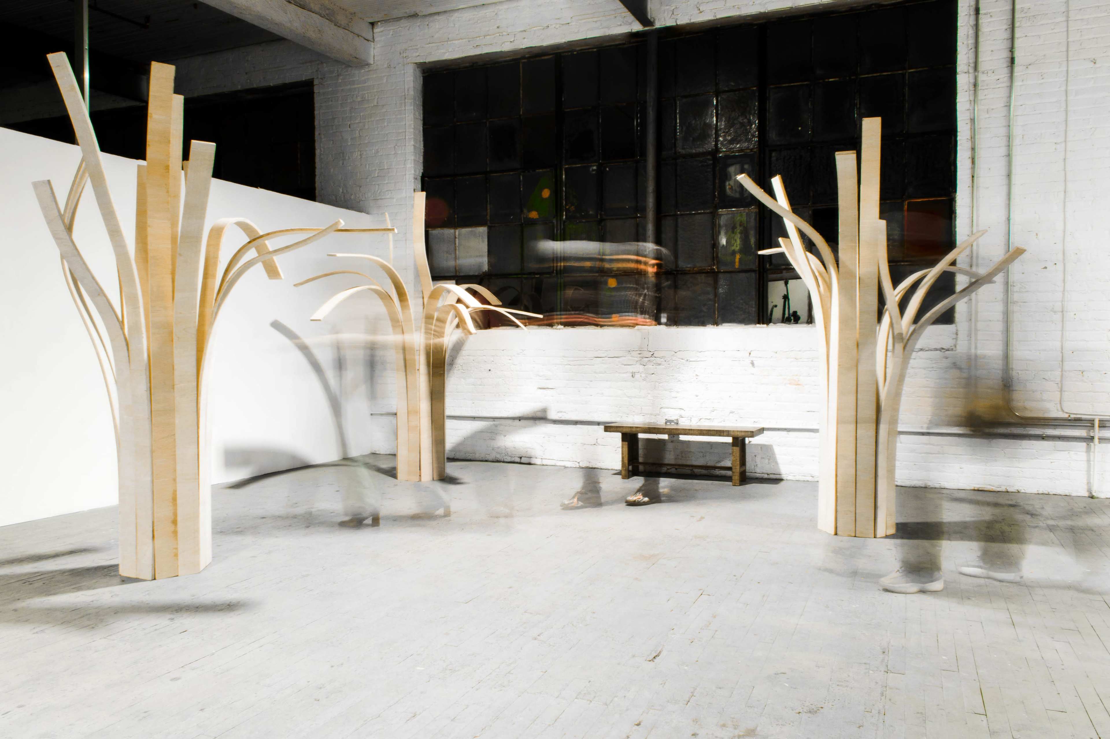 The image shows a spacious interior featuring three large wooden sculptures that resemble abstract trees, with a bench positioned in front of them.