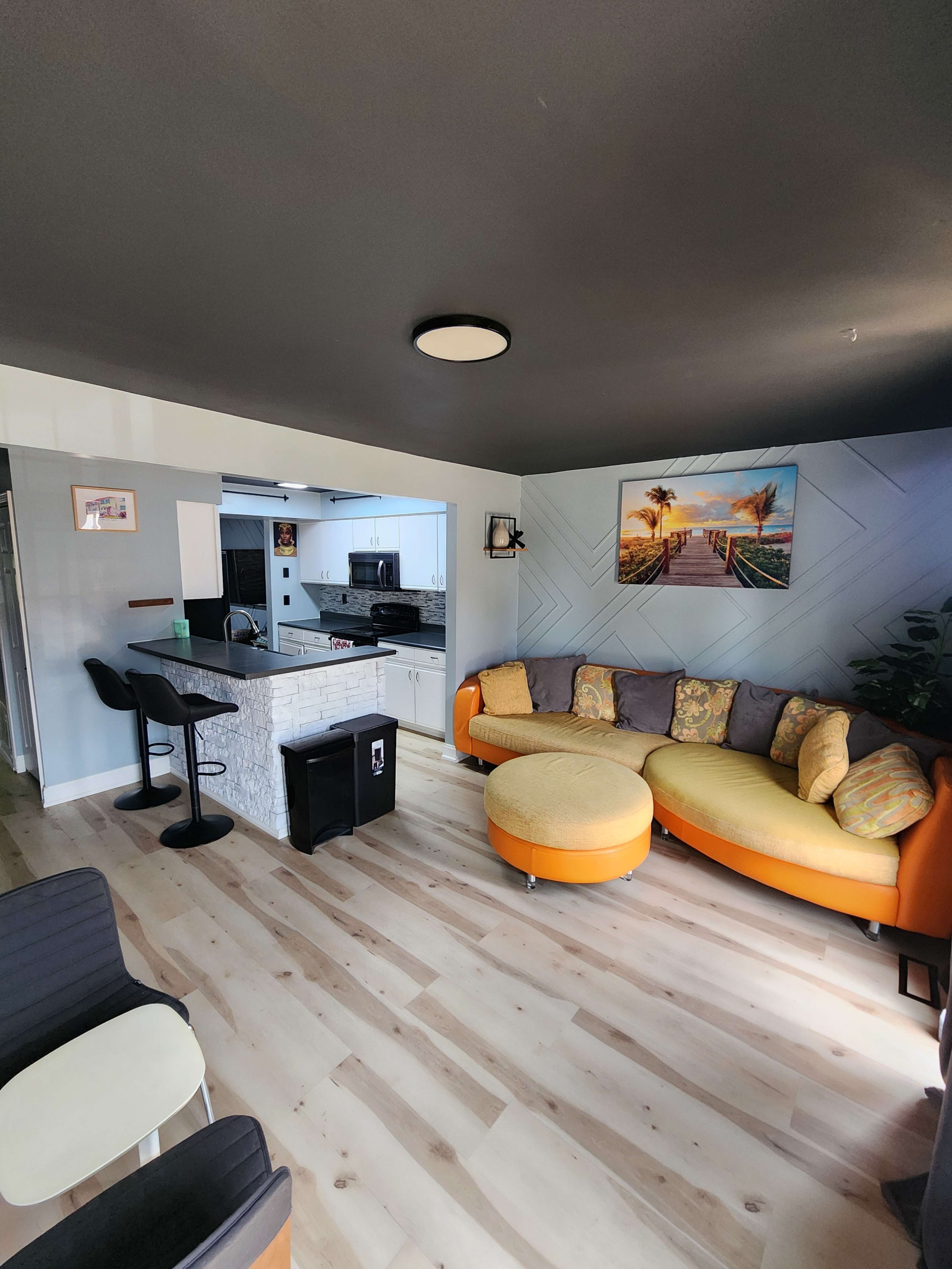 The image shows a living room with a circular orange sofa and a view of an adjacent kitchen area featuring a bar with two stools.