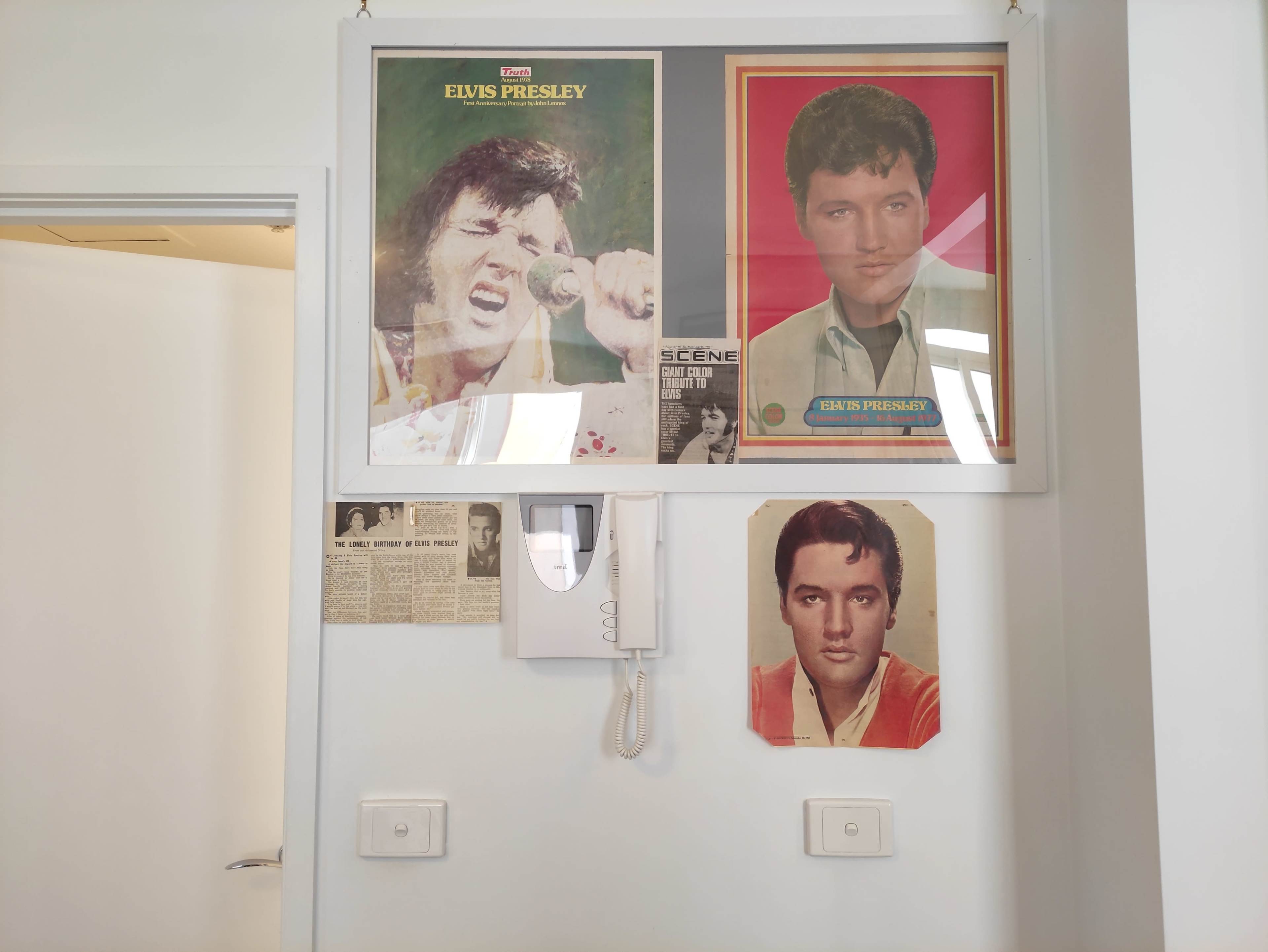 The wall features framed photos of Elvis Presley alongside an old newspaper clipping and a wall-mounted intercom.