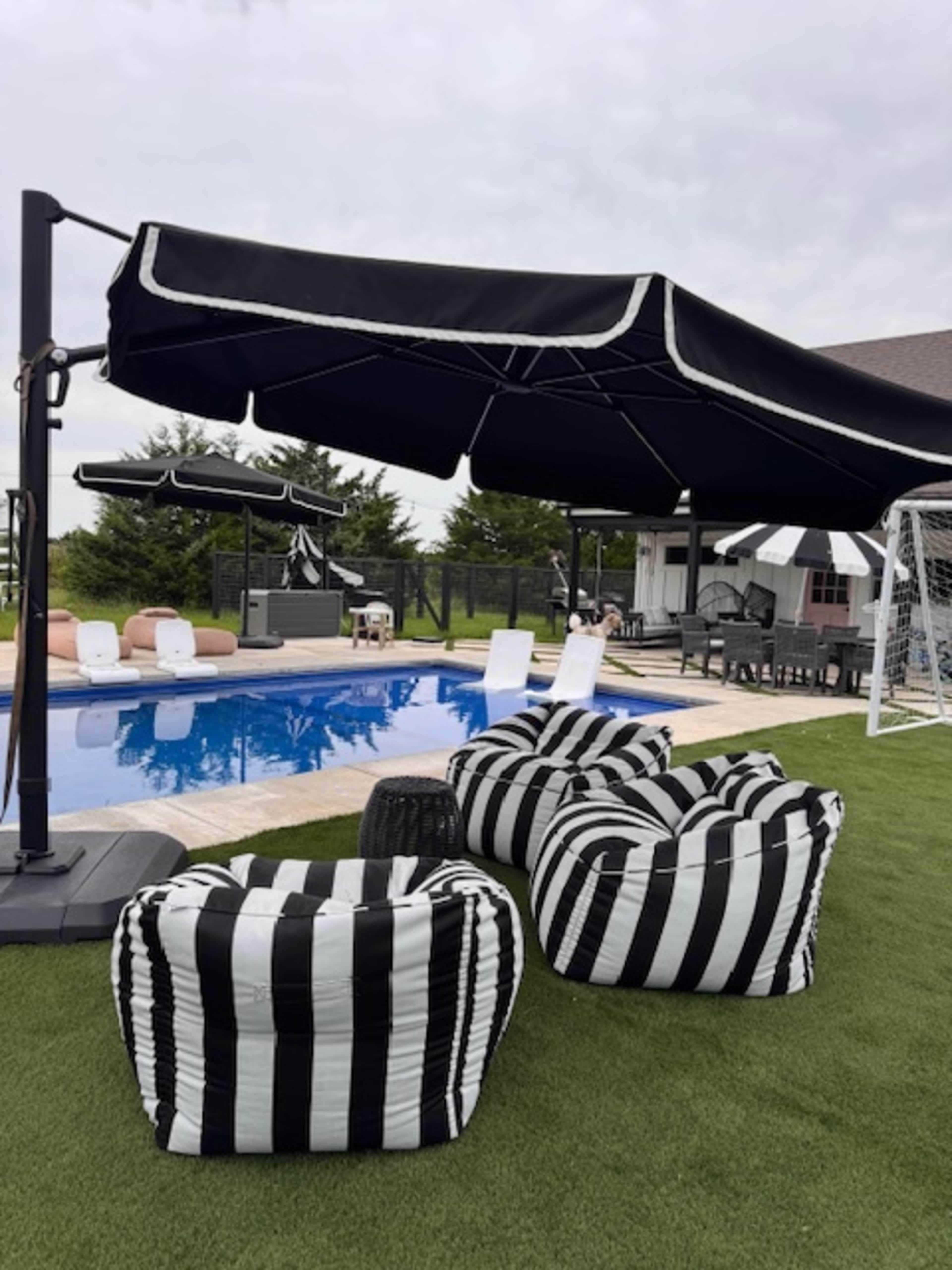 The image shows a poolside area with striped bean bag chairs, a large umbrella, and various seating options around a swimming pool.