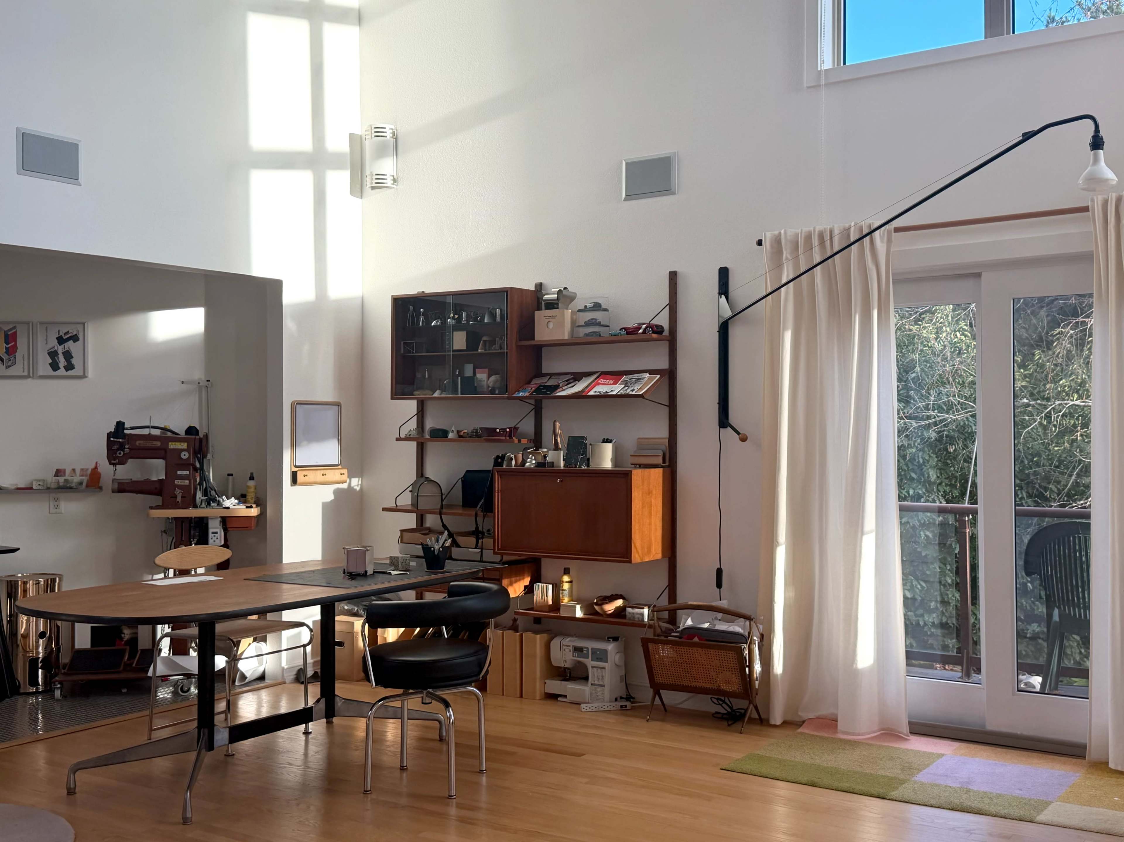 The image shows a well-lit modern interior featuring a wooden table with a black chair, a shelf filled with various items, and large windows opening to a view outside.