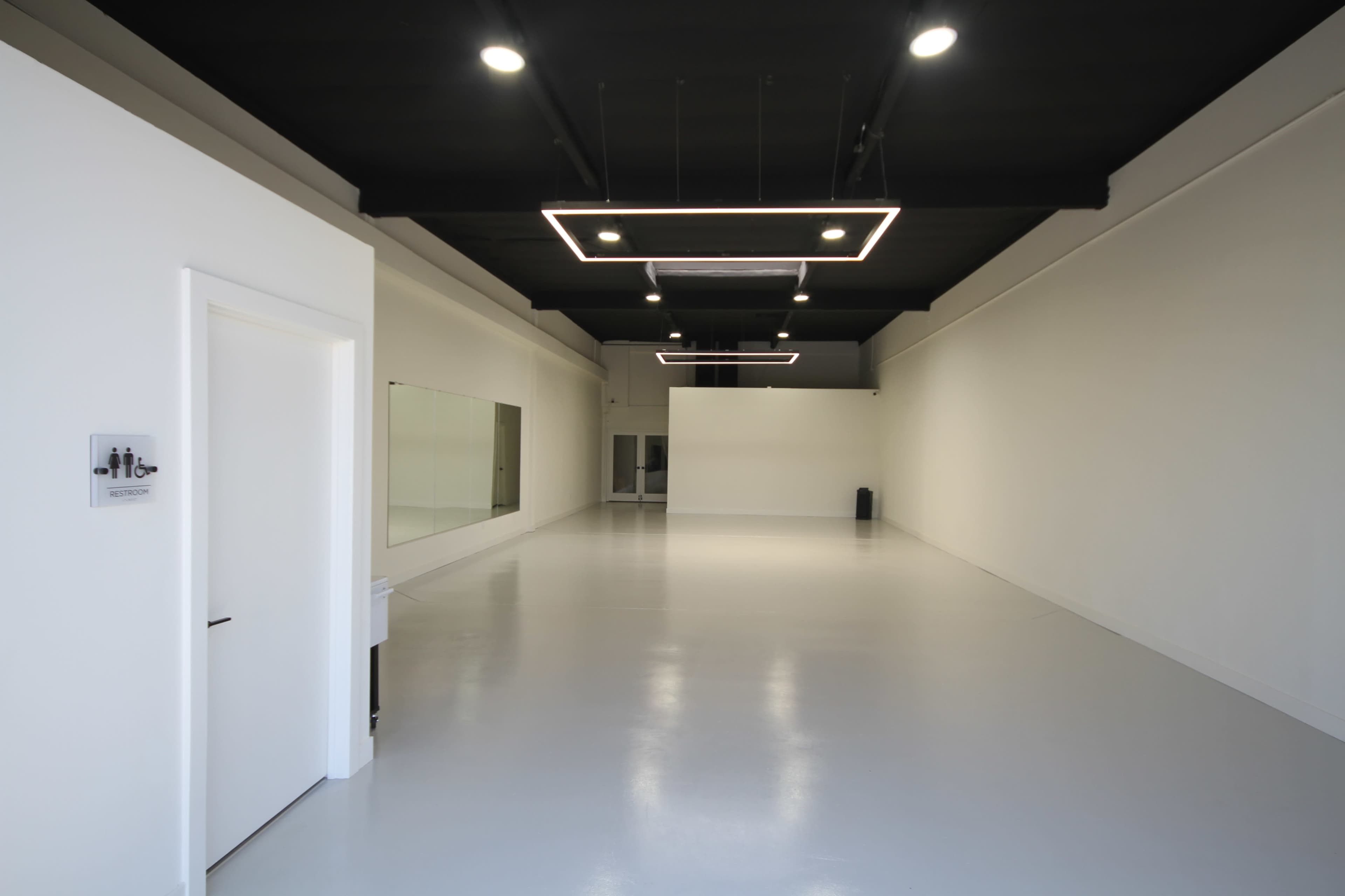 The image shows a spacious, empty room with white walls, a polished floor, and modern lighting fixtures overhead.