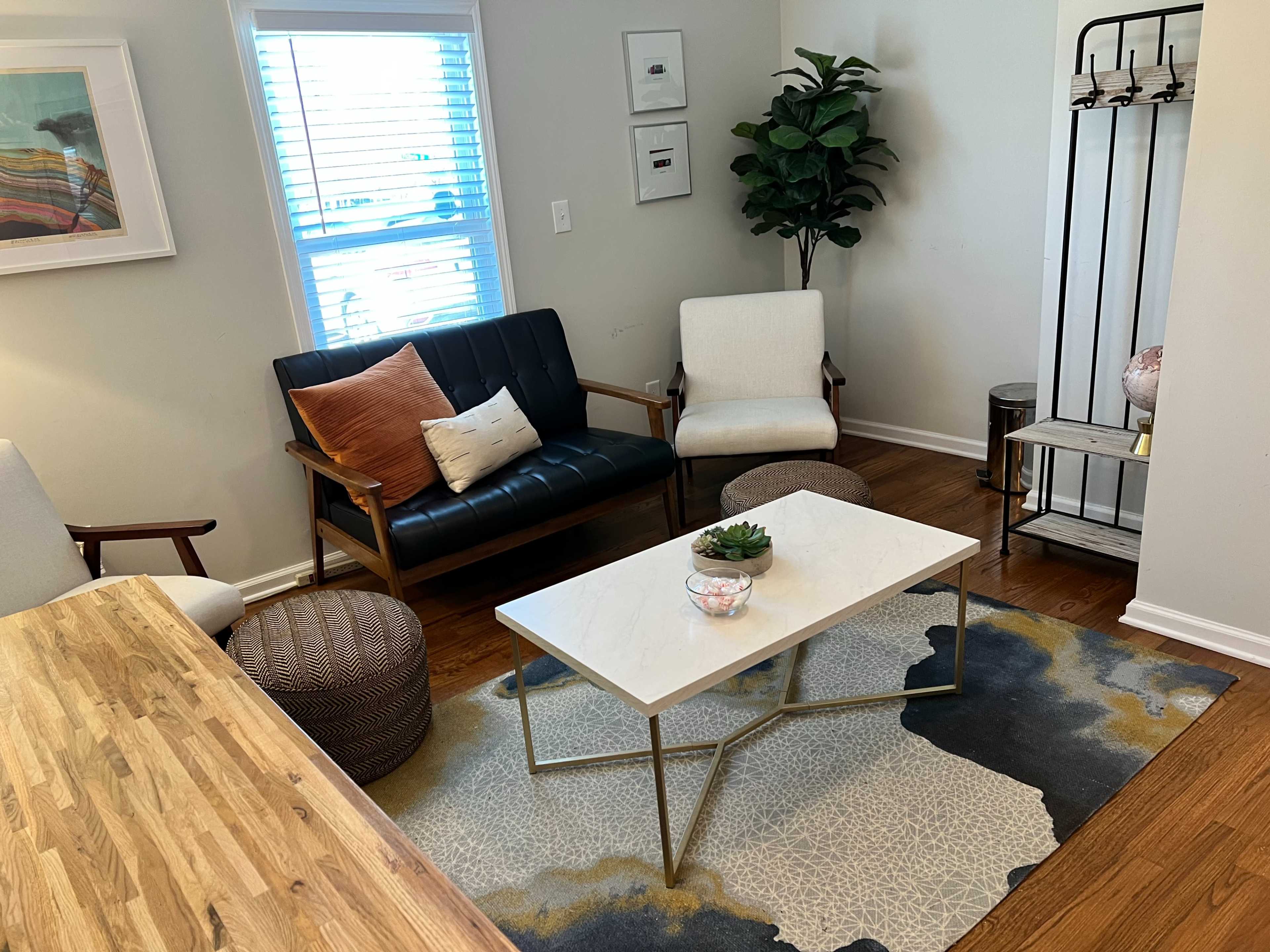 The image shows a cozy living room with a coffee table in the center, surrounded by a dark leather sofa, a light-colored armchair, a potted plant, and a decorative rack.