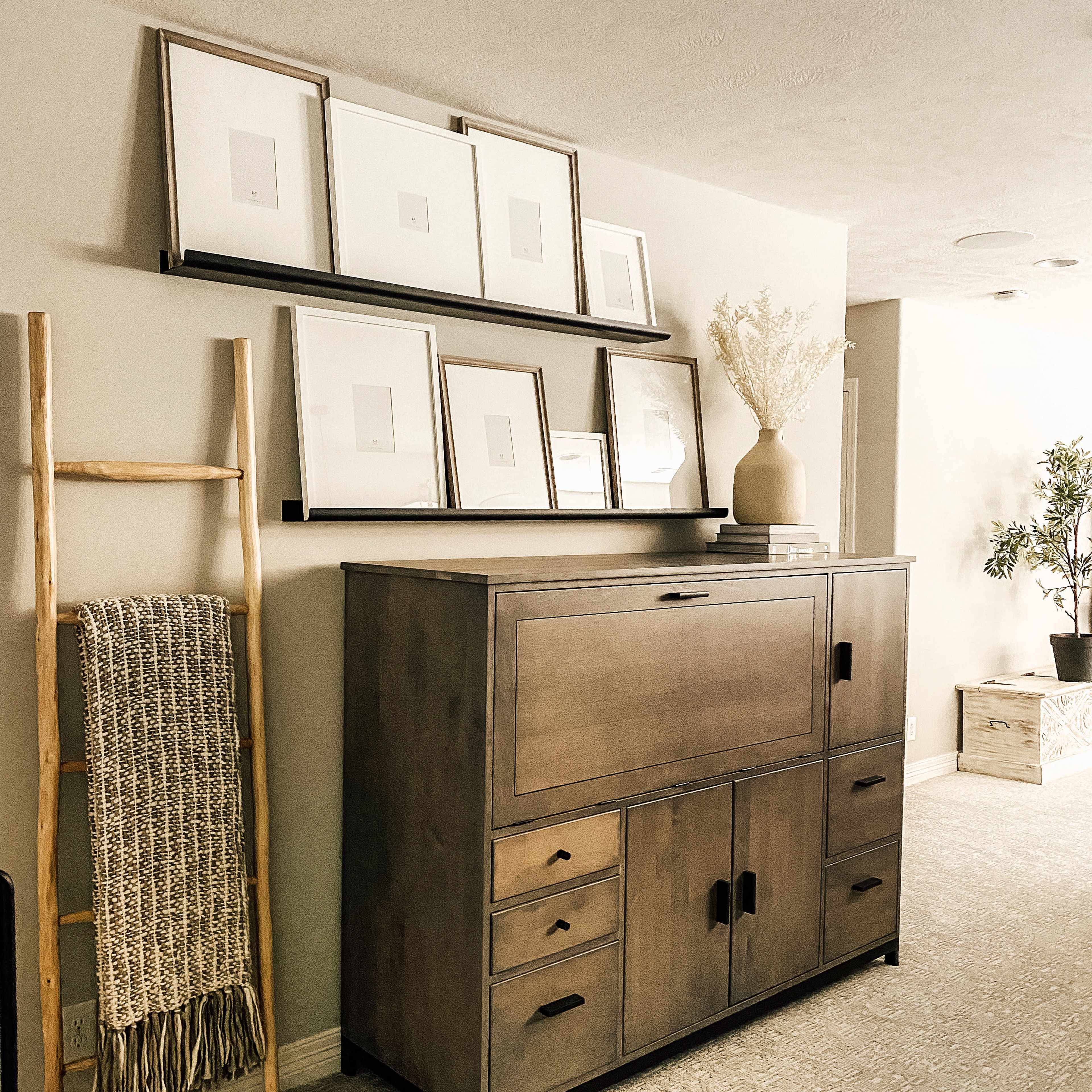 A wooden cabinet with multiple drawers and a drop-down door sits beneath a shelf displaying empty frames and a vase, next to a woven ladder and a potted plant.