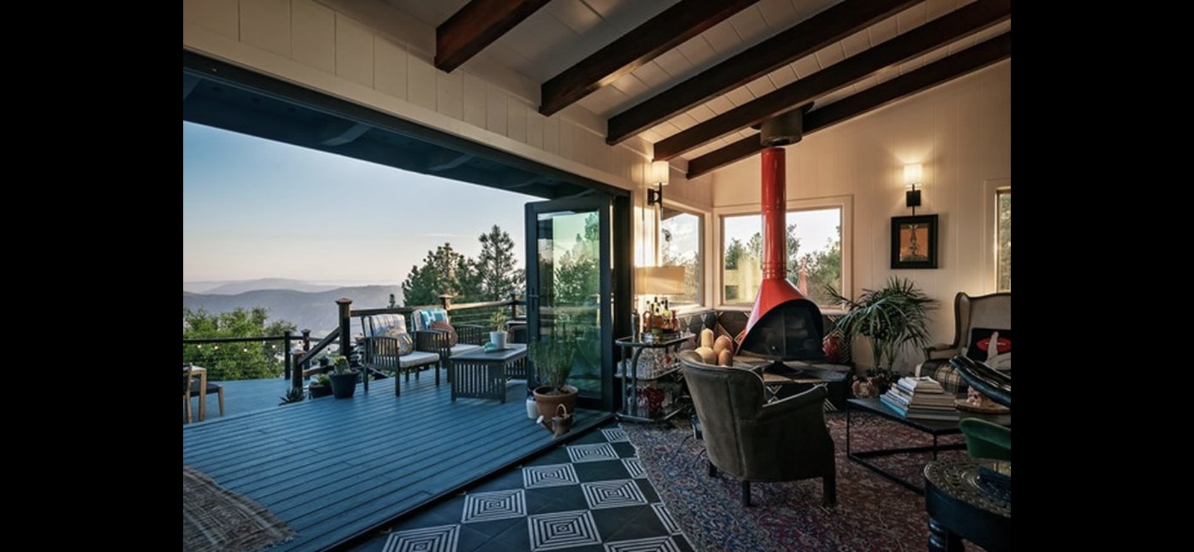 A modern living room with large glass doors opening to a scenic view, featuring a red fireplace, comfortable seating, and a decorative rug.