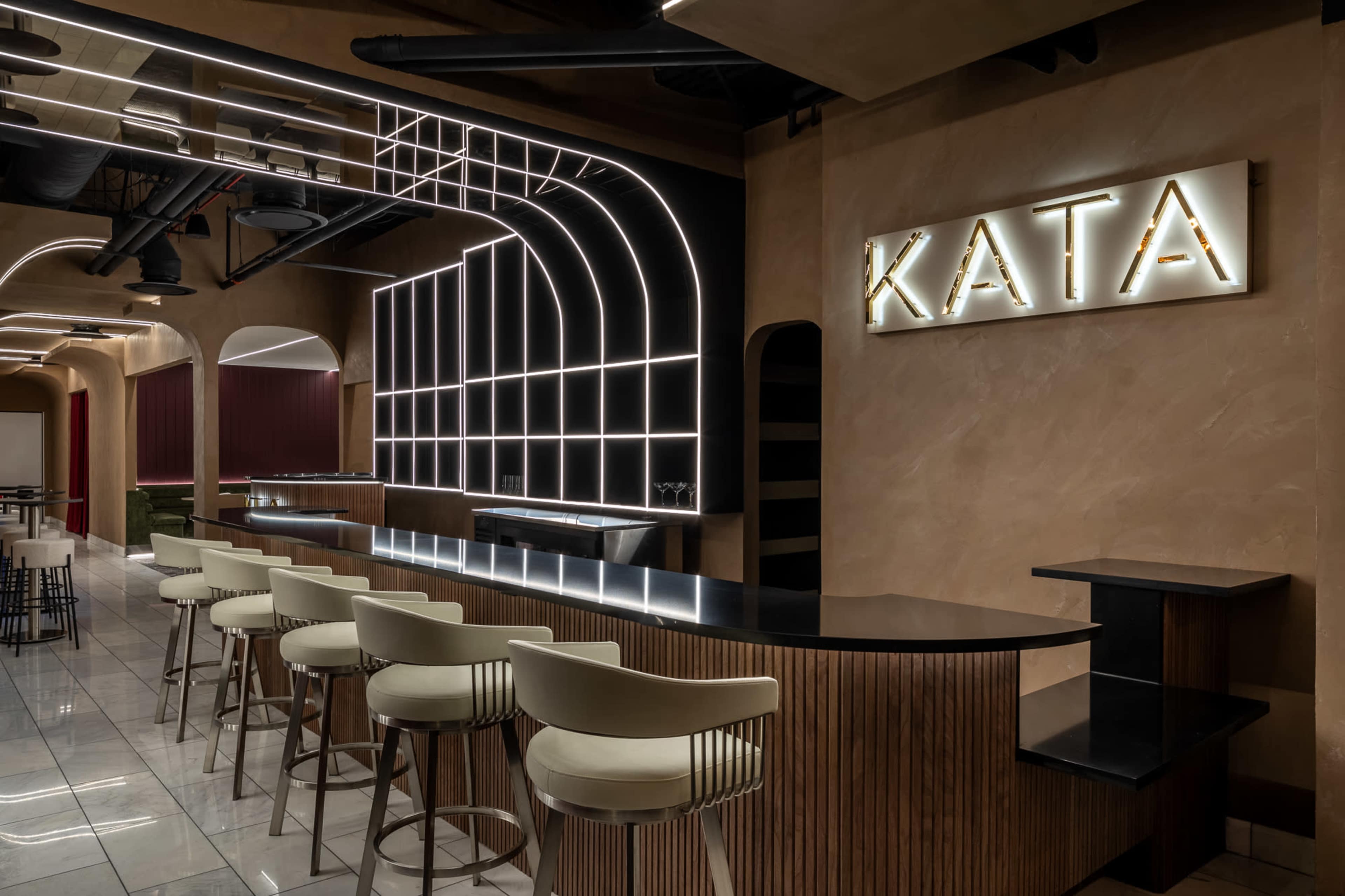 The image shows a modern bar interior with a sleek black countertop, white bar stools, and a prominent illuminated sign that reads "KATA."