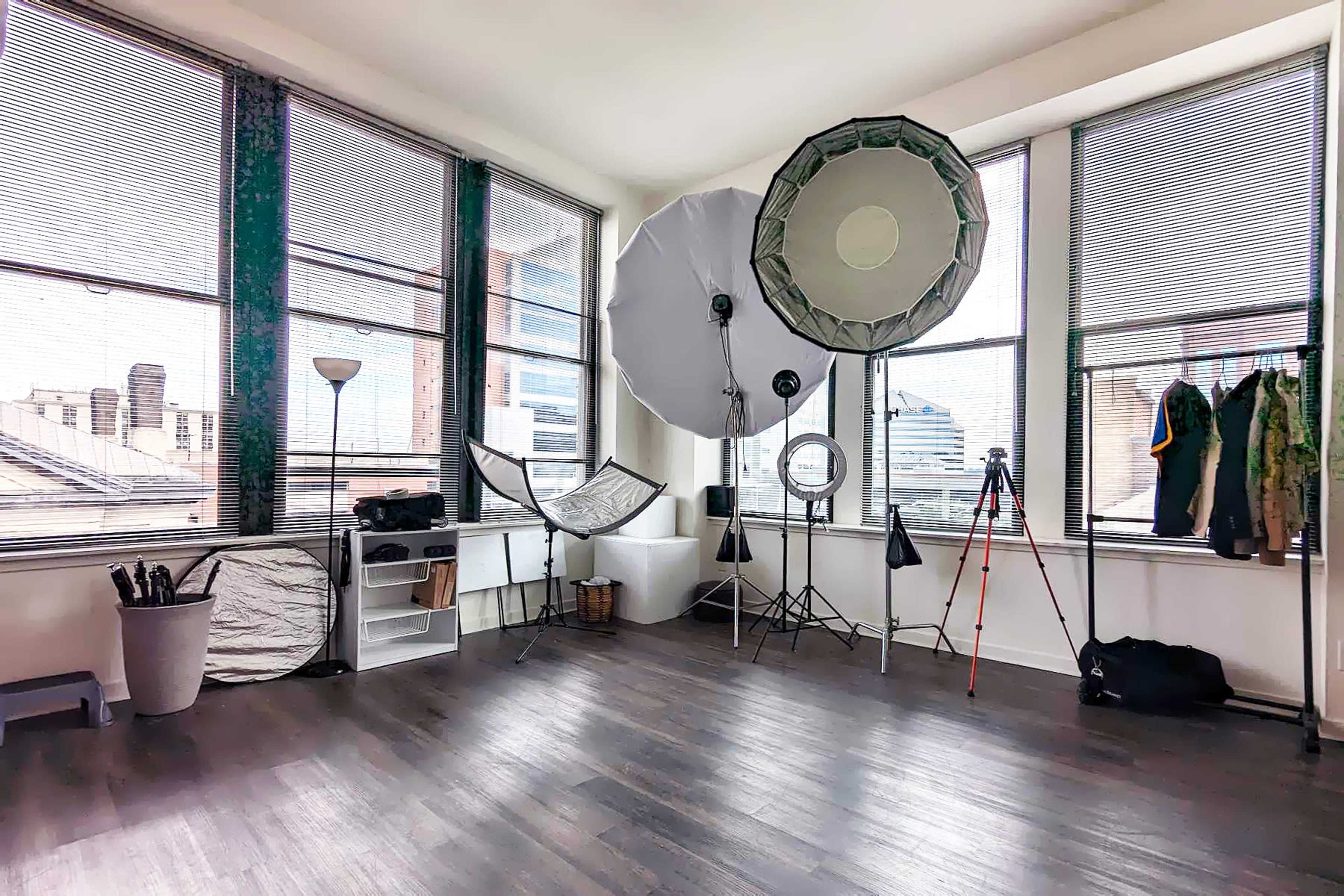 A spacious room with large windows, photography equipment including softboxes and tripods, and various items hanging on hooks.