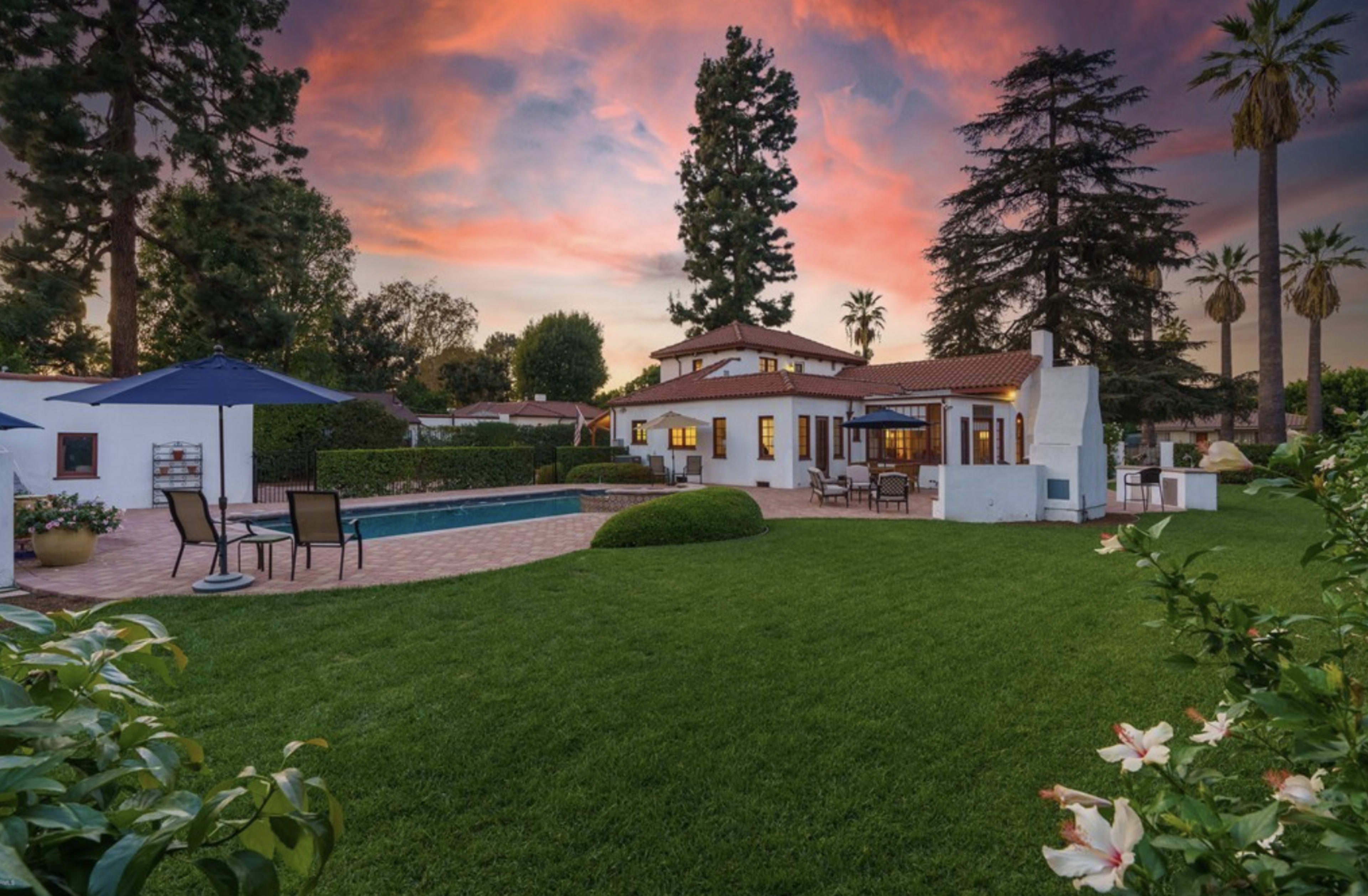 A backyard featuring a swimming pool, lush green grass, and a white house with red-tiled roofing, set against a vibrant sunset sky.