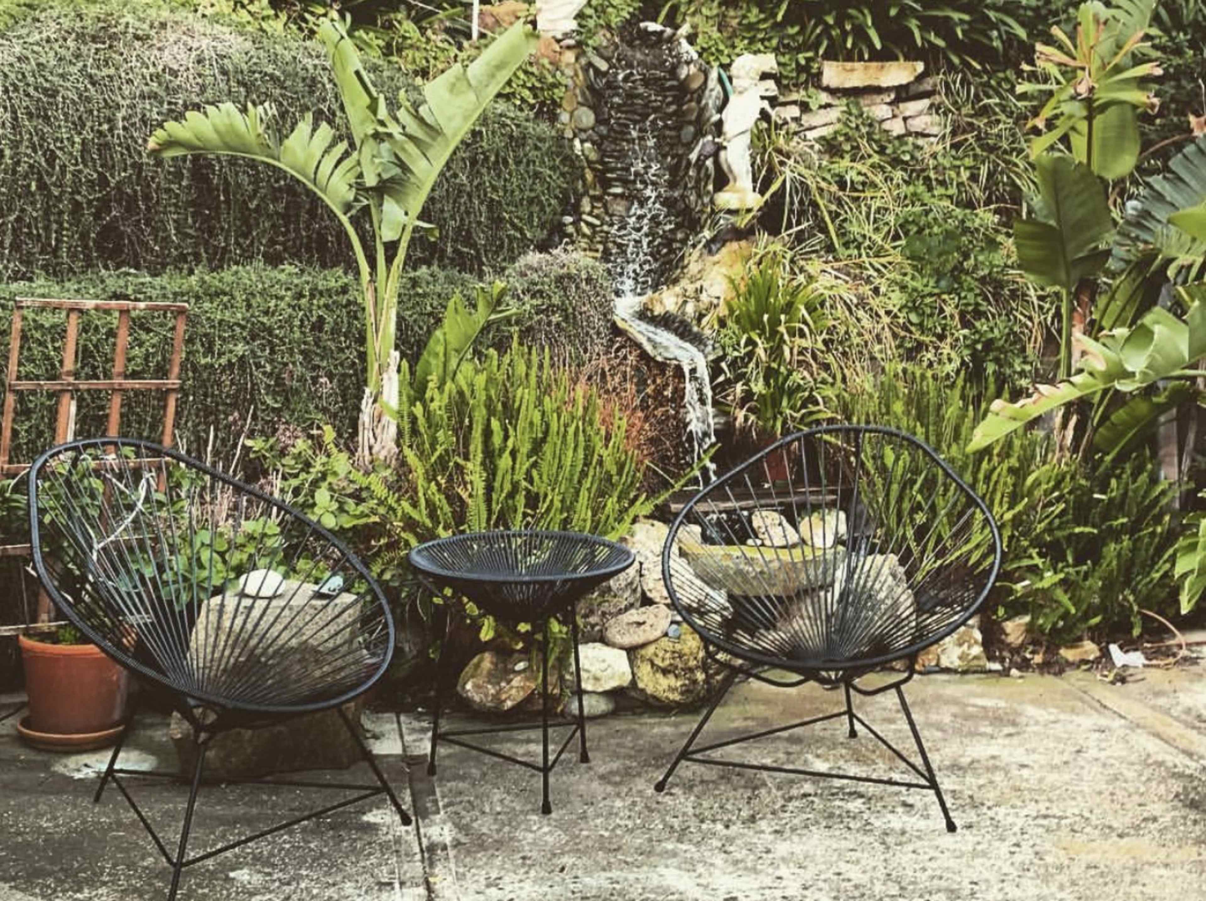 Two black wire chairs and a small round table arranged in a garden with a waterfall and lush greenery.