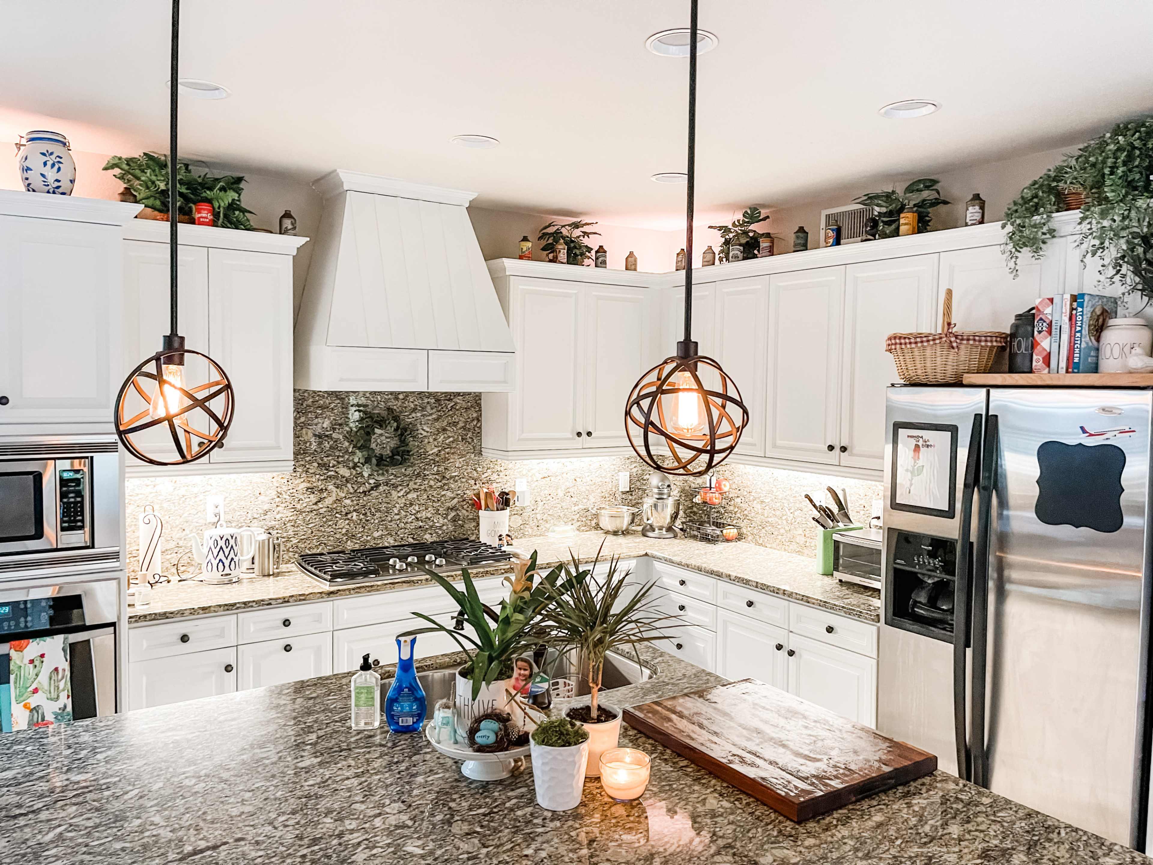 A modern kitchen features white cabinets, granite countertops, and pendant lights, with various plants and kitchen accessories displayed throughout.