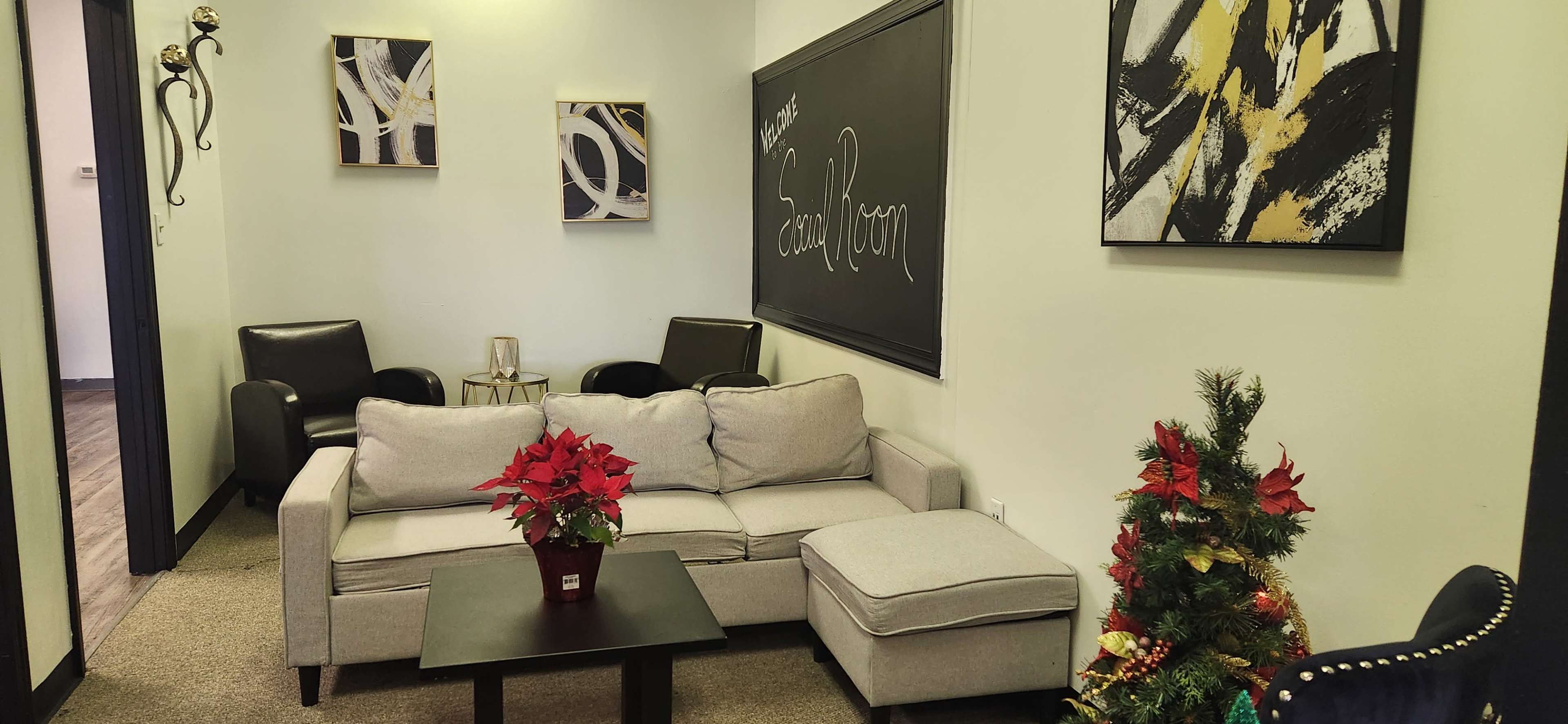 The image shows a waiting room with a gray couch, a small black table, and two black armchairs, decorated with artwork and a small festive plant.