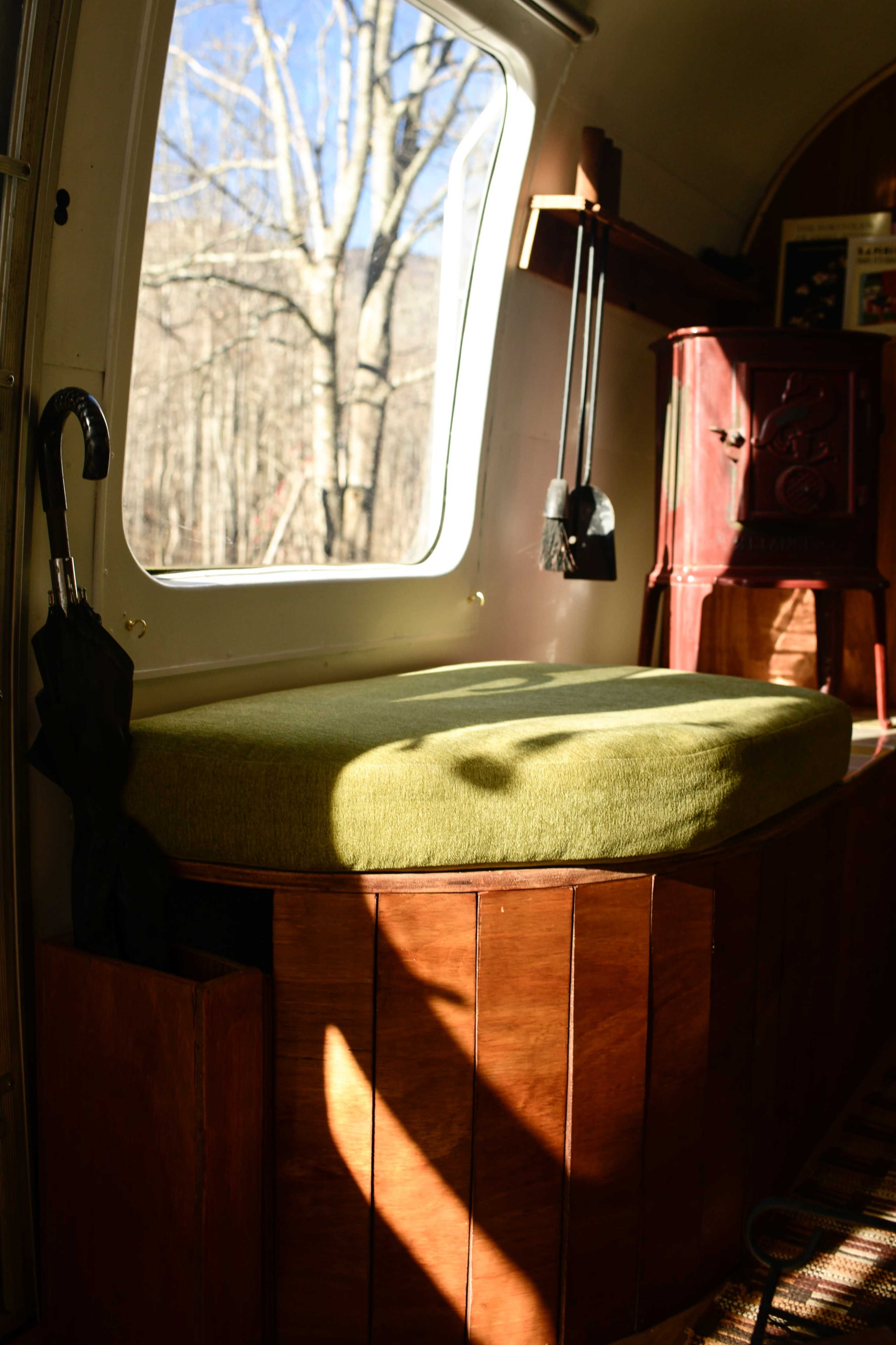 A green cushioned bench sits beside a large window, offering a view of trees outside, with an umbrella holder attached to the side.