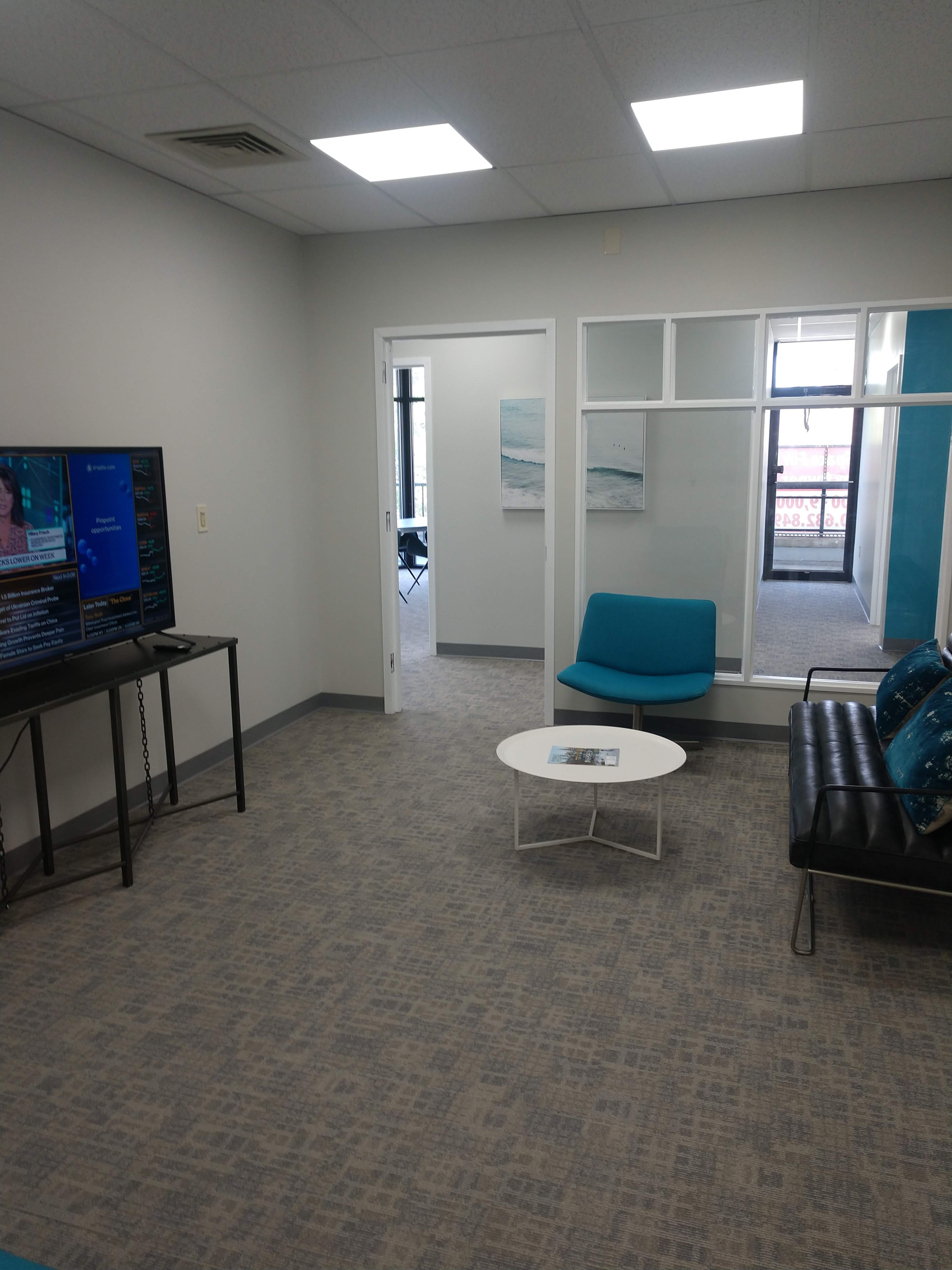 The image shows a modern waiting area with a television, a blue couch, and a round coffee table, featuring light gray walls and carpet.