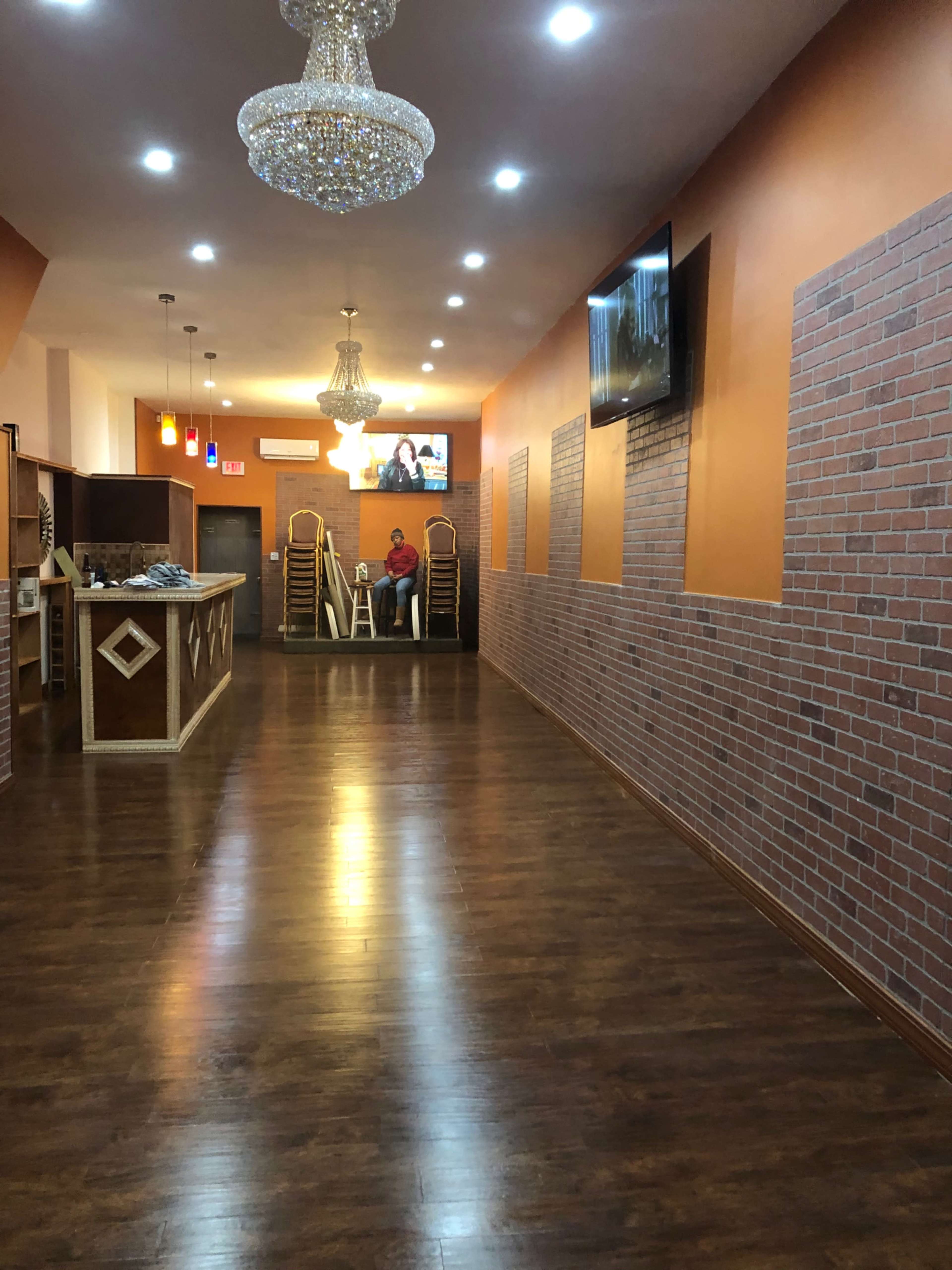 A long, well-lit hallway with wooden flooring and brick-patterned walls, featuring a chandelier and a television mounted on one wall.