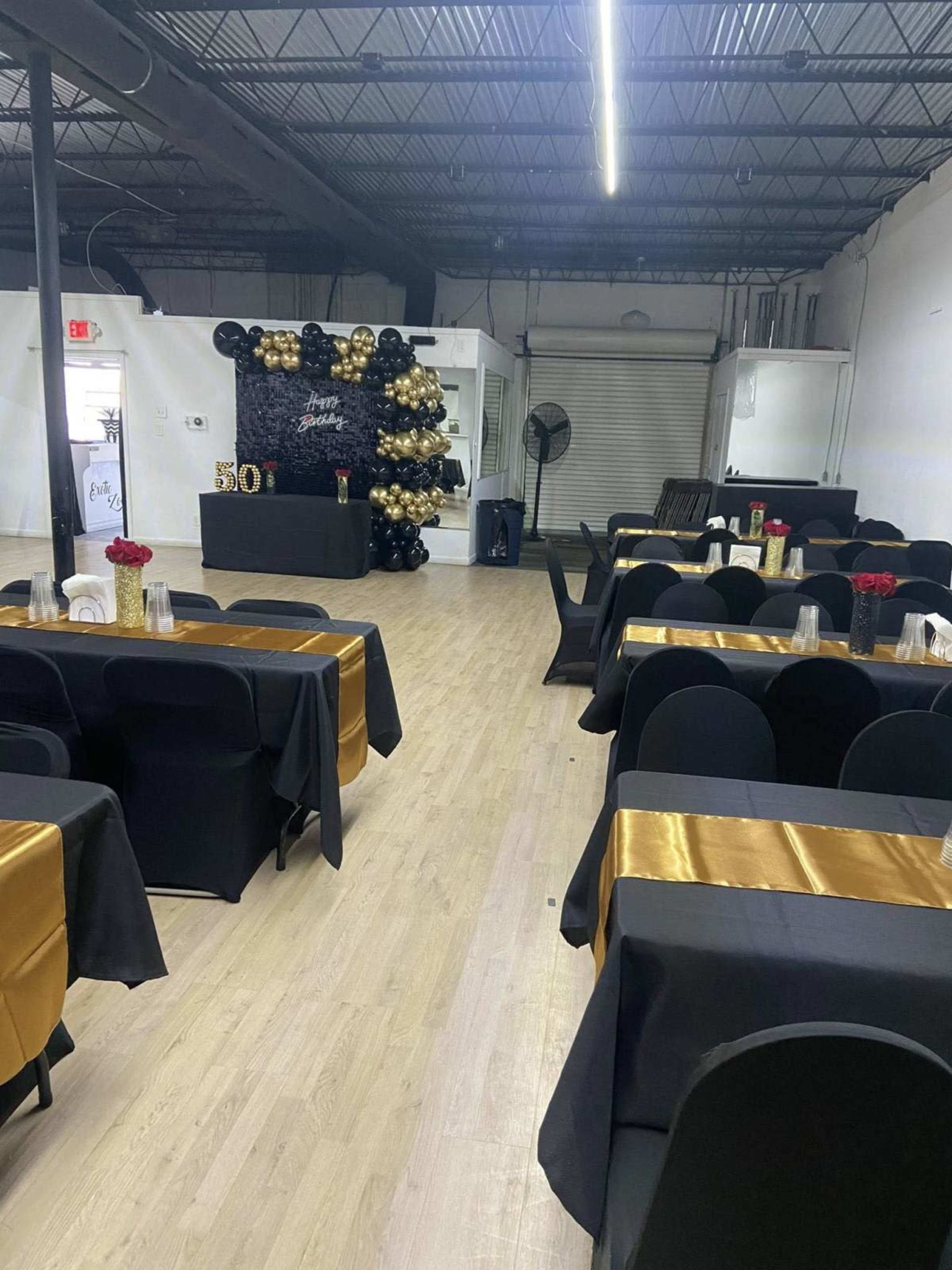 The image shows an event space set up for a celebration, featuring black and gold tables, a backdrop decorated with balloons, and a stylish arrangement for seating.