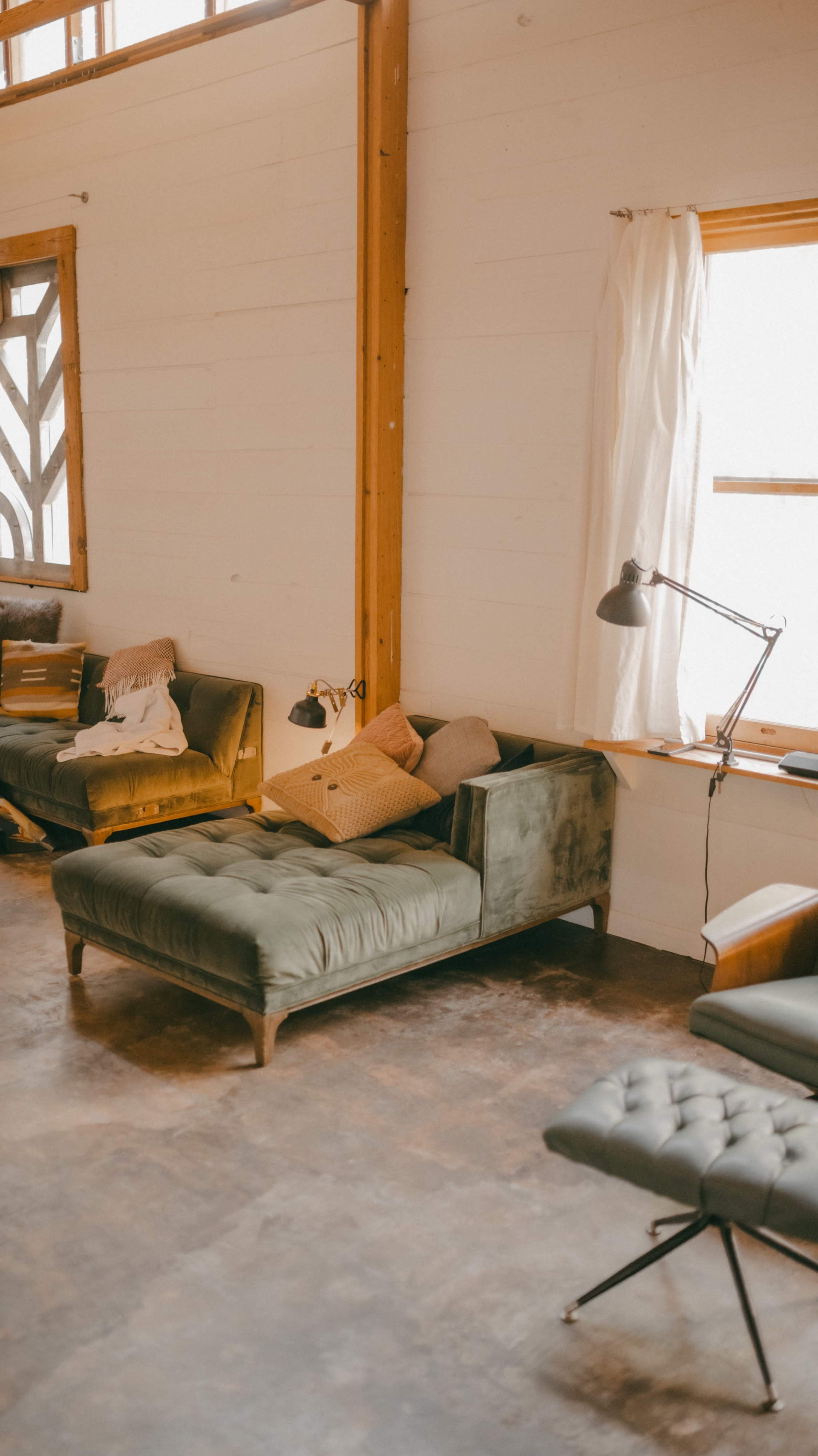 The image shows a minimalist interior with green sofas, a floor lamp, and large windows allowing natural light.