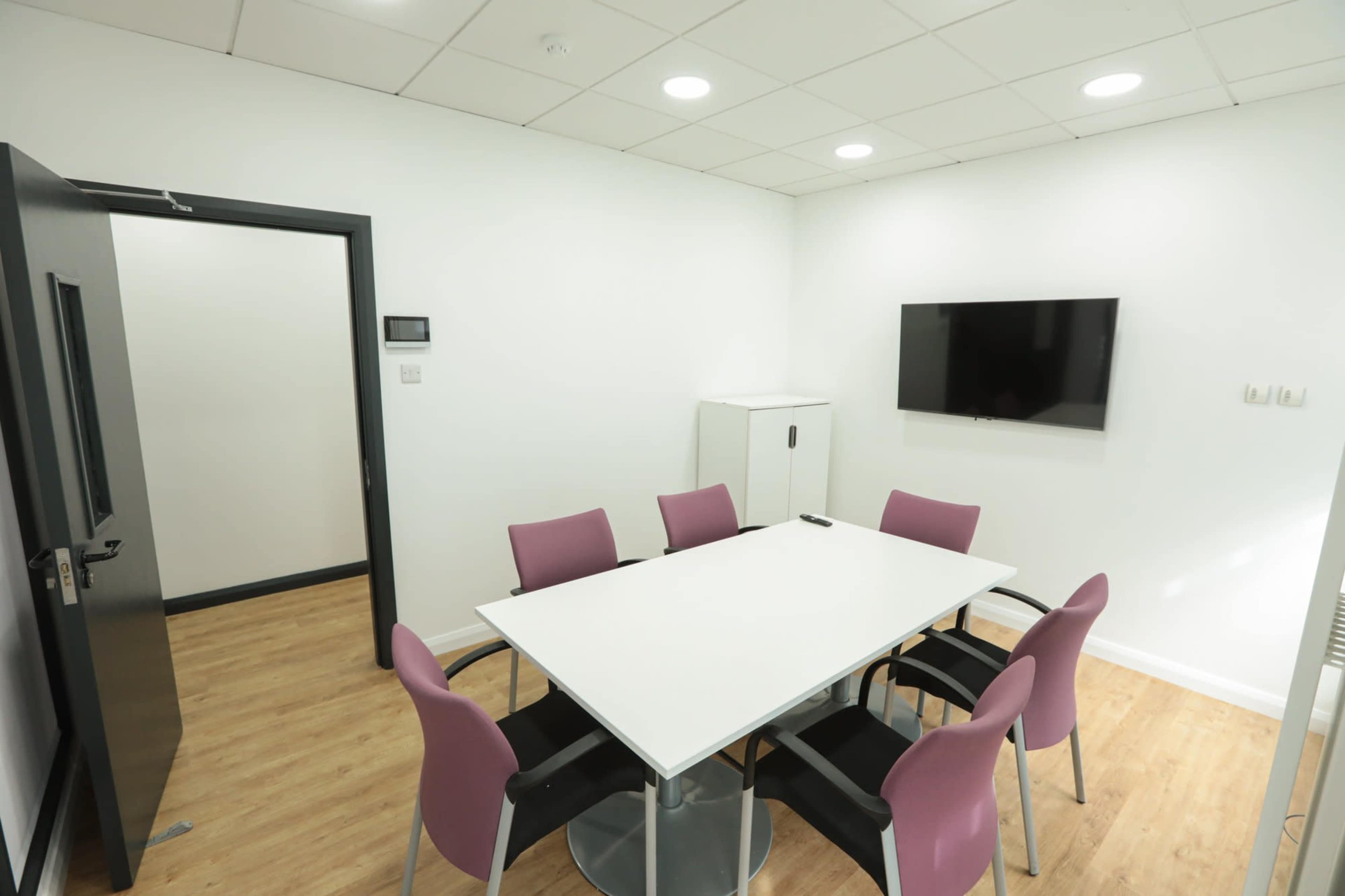 A small conference room features a round table surrounded by six purple chairs, a wall-mounted television, and a cabinet, all within a bright, minimalist space.