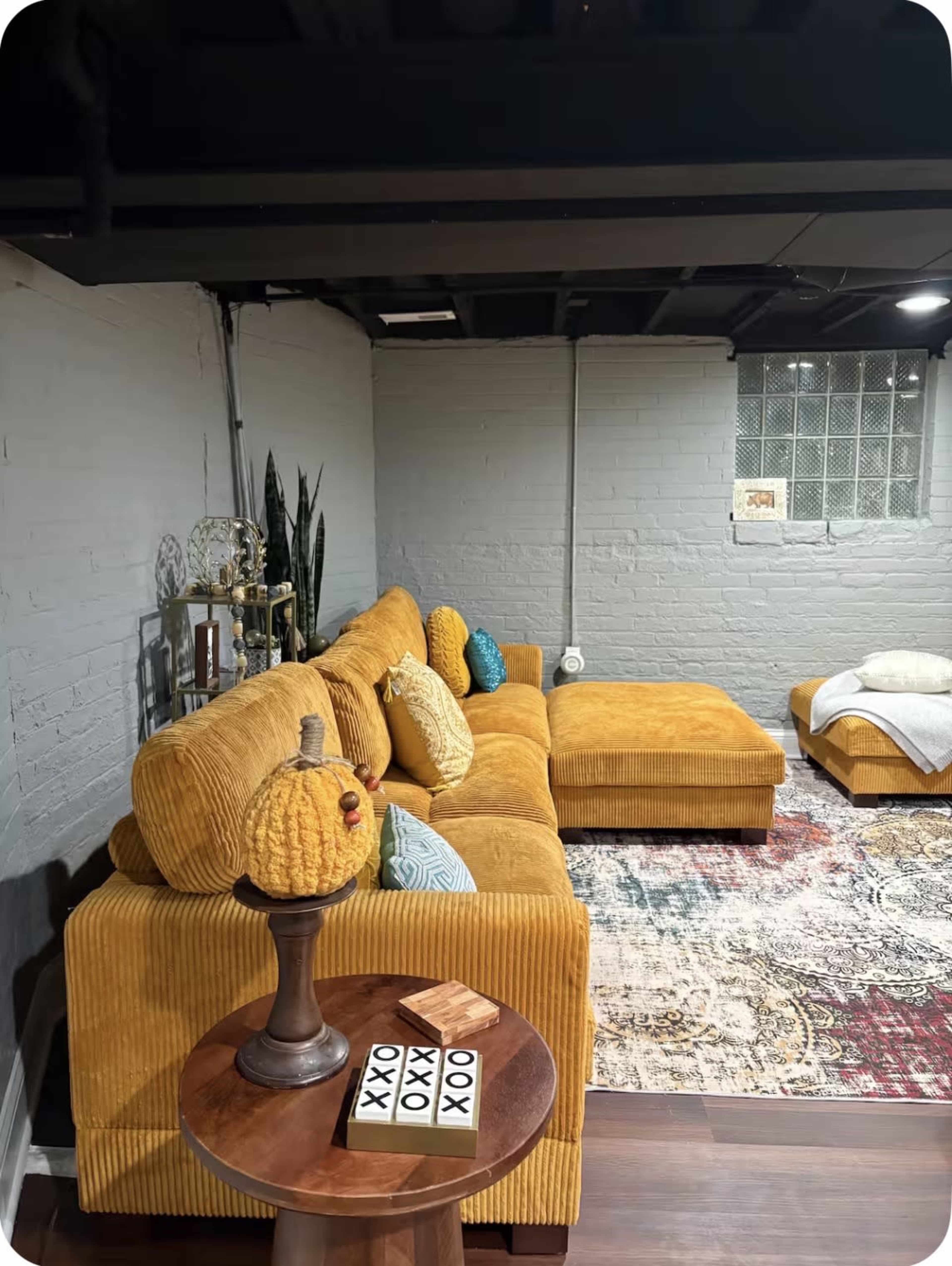 The image shows a cozy yellow corduroy sectional sofa accompanied by a small round table, a decorative pumpkin, and a colorful rug in a well-lit basement living area.