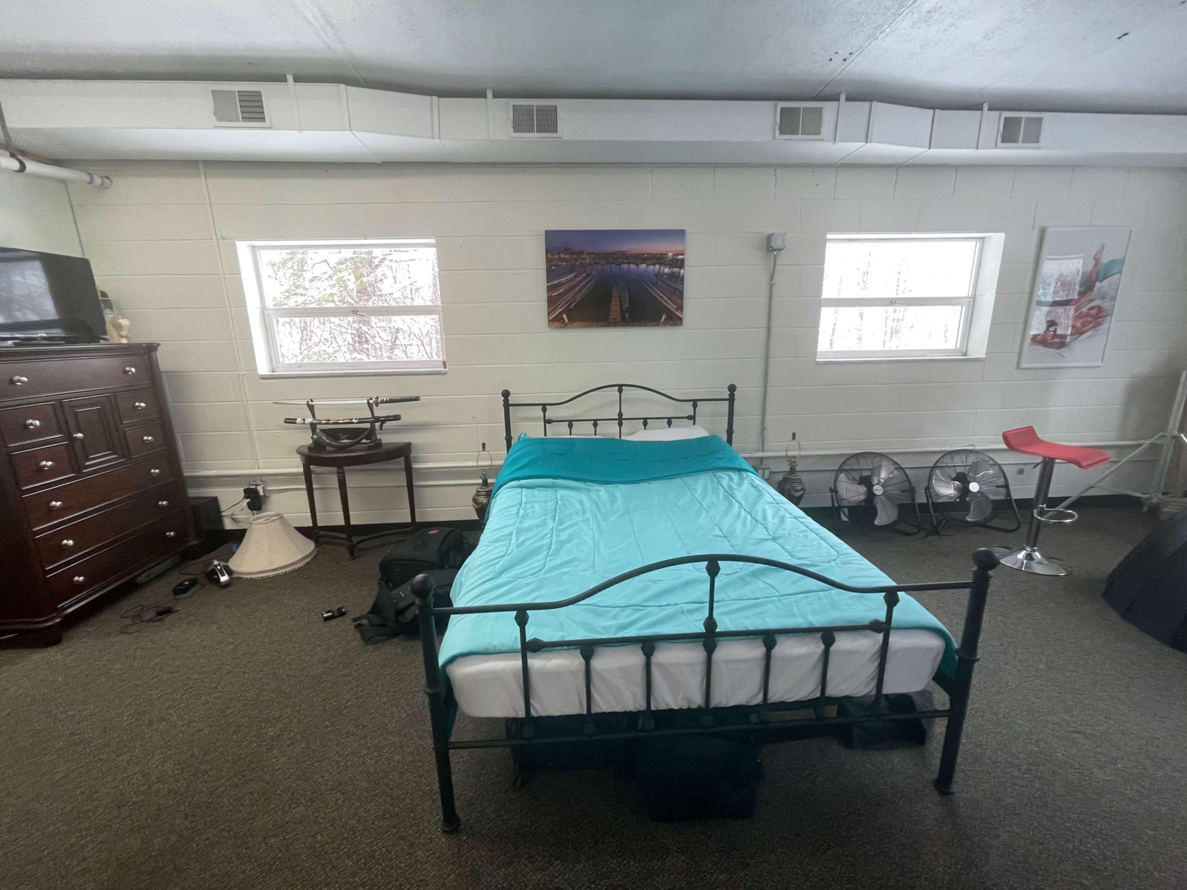 A simple bedroom with a bed featuring a light blue comforter, a dresser, two windows, and various items scattered on the floor, including fans and a lamp.