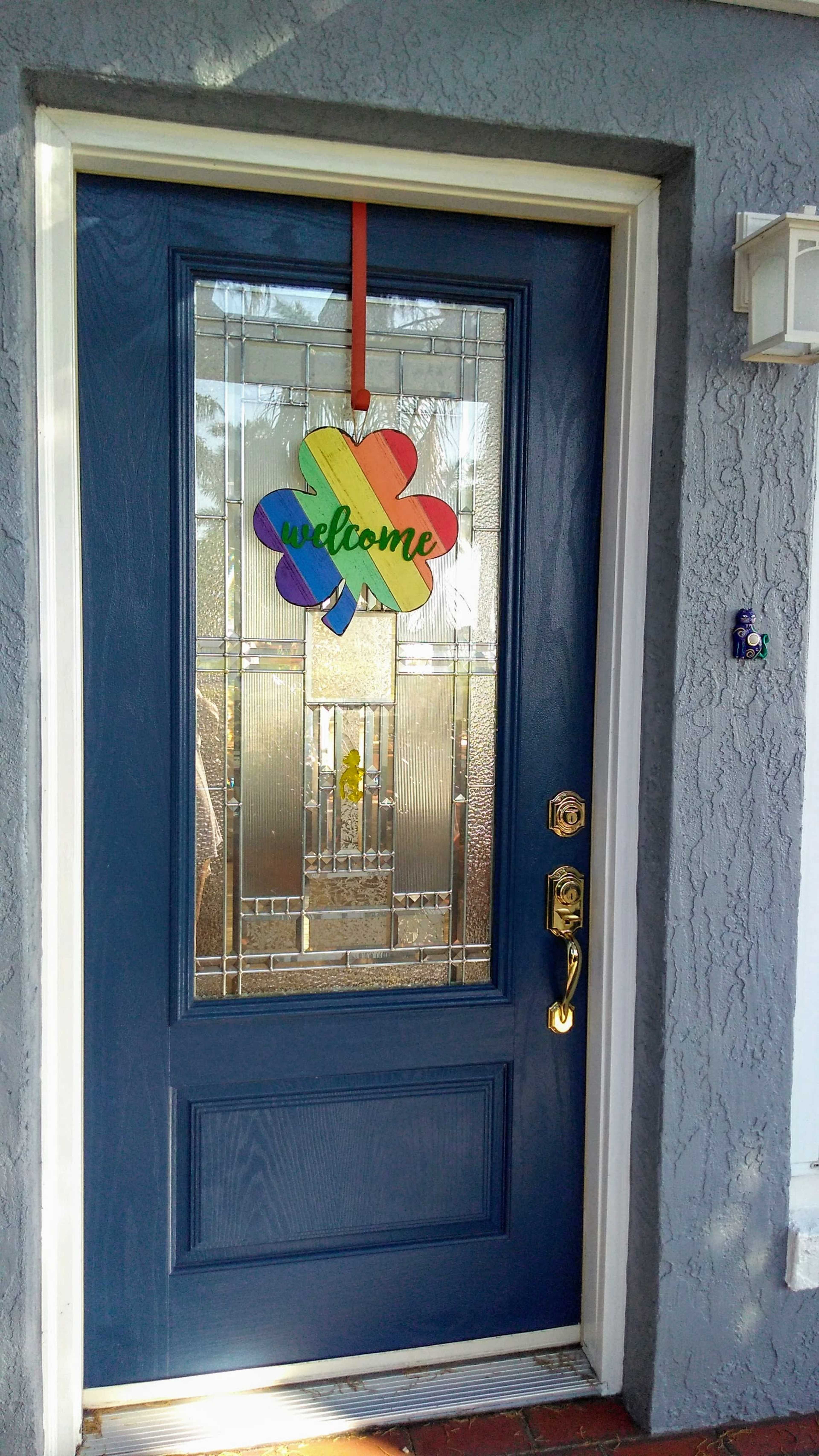 The image shows a blue front door with a colorful "welcome" sign shaped like a flower hanging from the top.