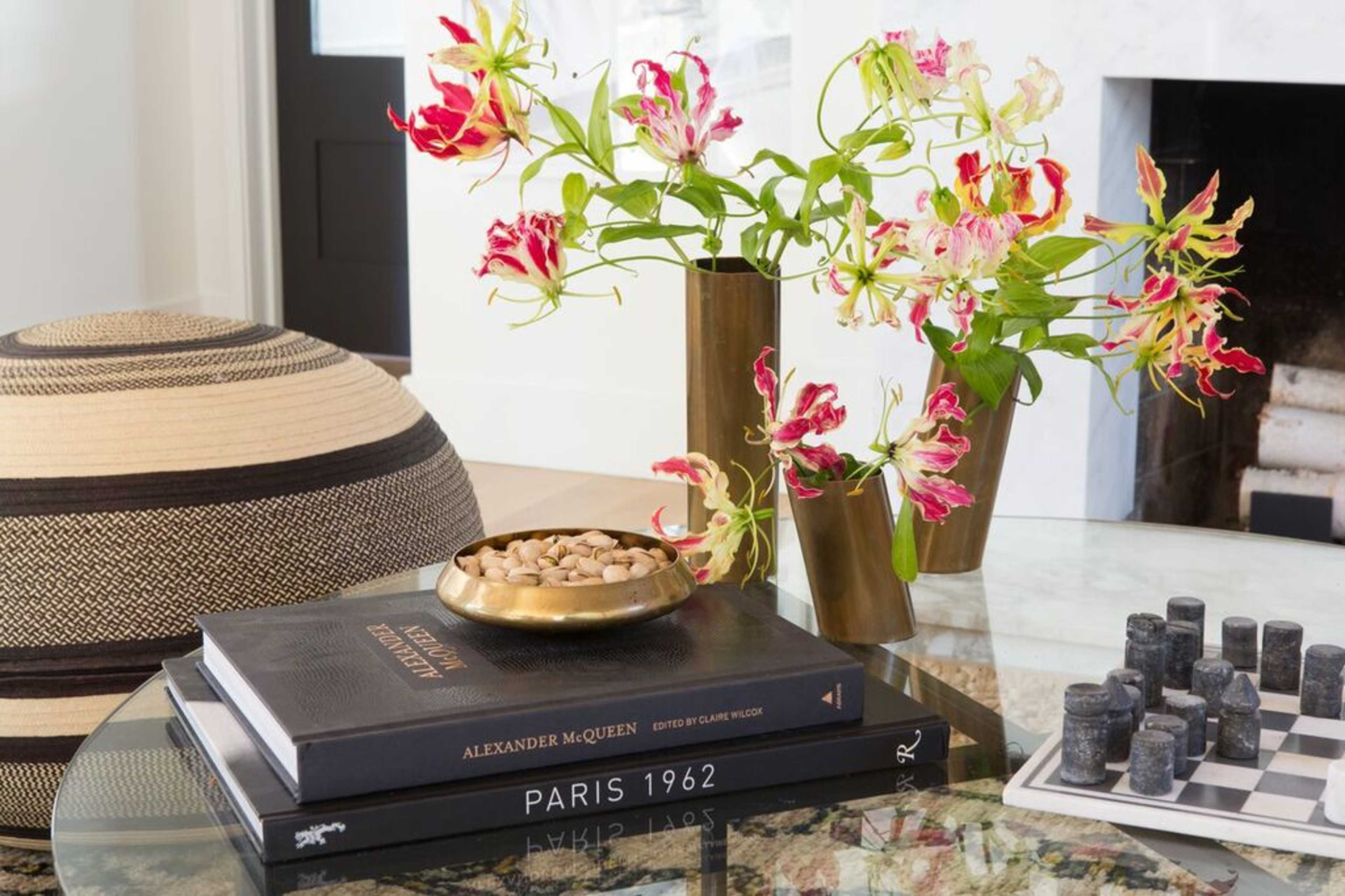 A glass table displays several stacked books, a decorative bowl with pebbles, and vibrant flowers in brass vases, with a woven ottoman and a fireplace in the background.
