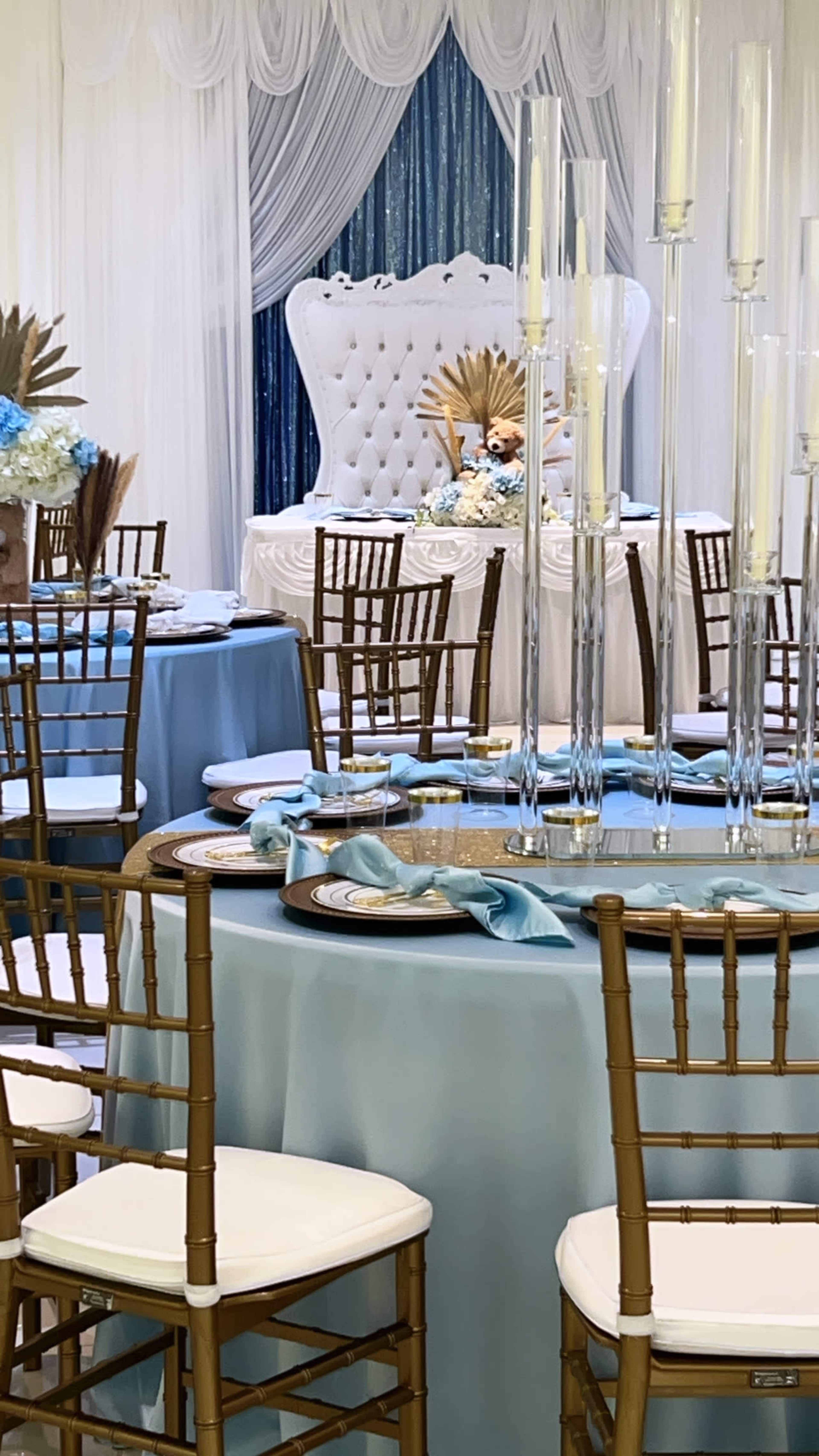 A decorated banquet hall features elegantly set tables with light blue tablecloths, gold chiavari chairs, and a backdrop of a white throne surrounded by floral arrangements and tall candle holders.