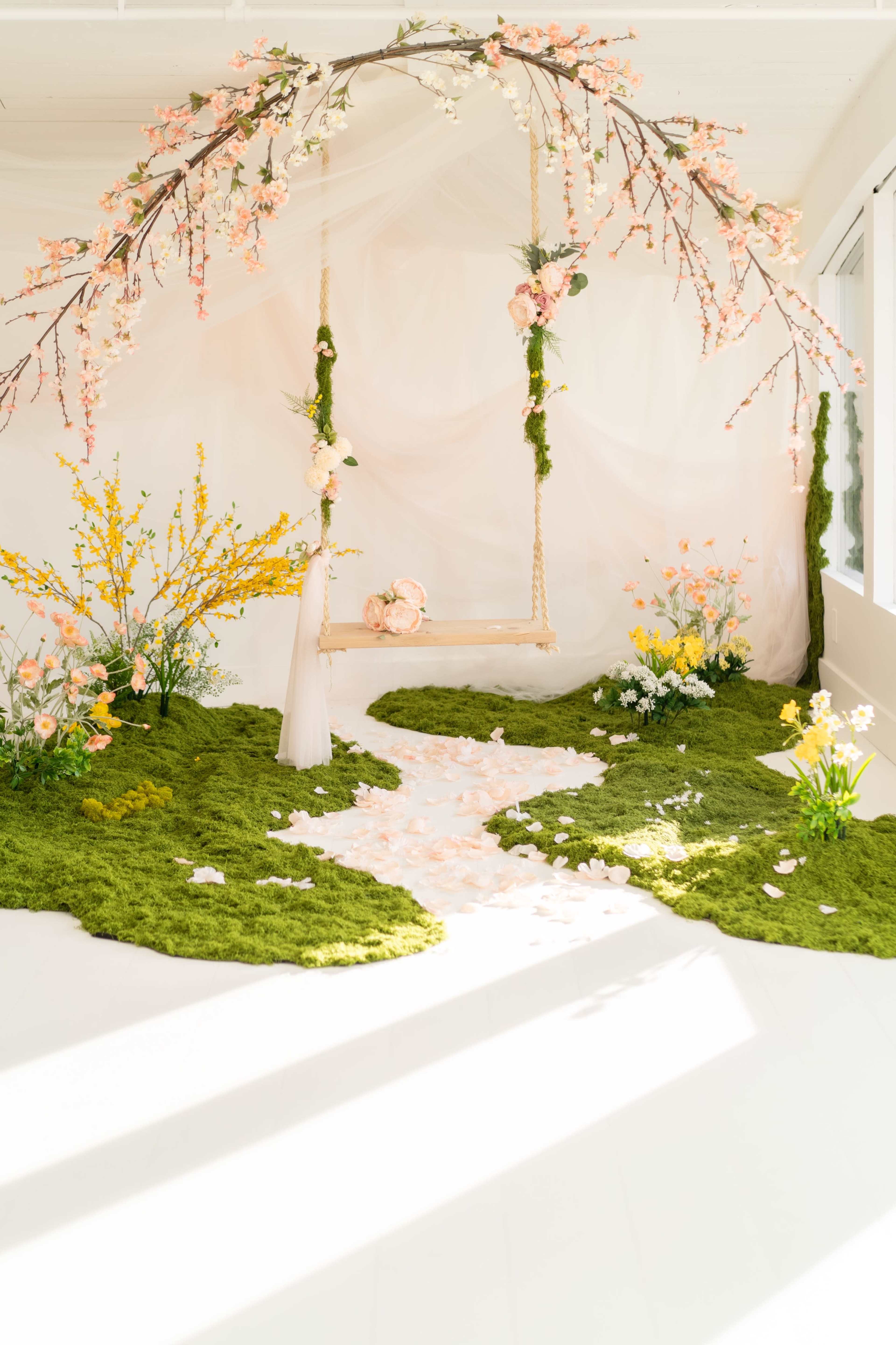 The image shows a beautifully decorated indoor space featuring a swing adorned with flowers, surrounded by greenery and floral arrangements.