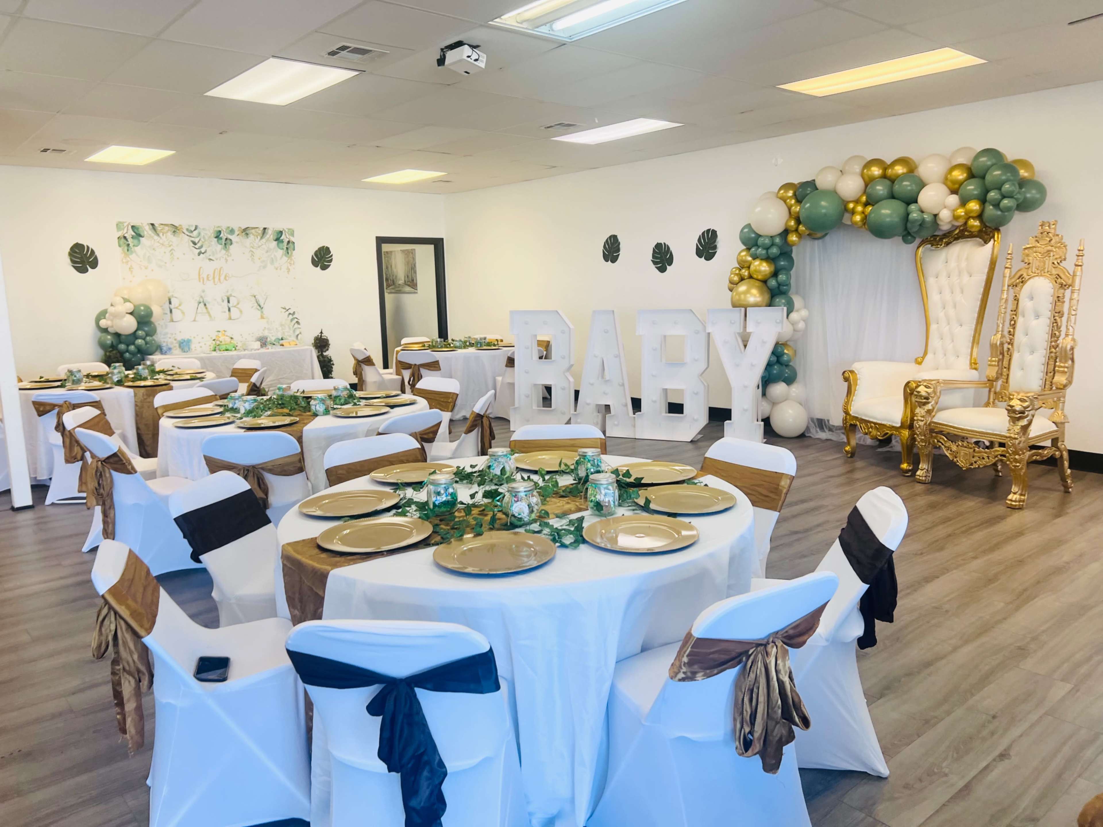 A decorated event space with several round tables set for a celebration, featuring gold plates, white and gold chairs, and a large "BABY" display along with balloon decorations.