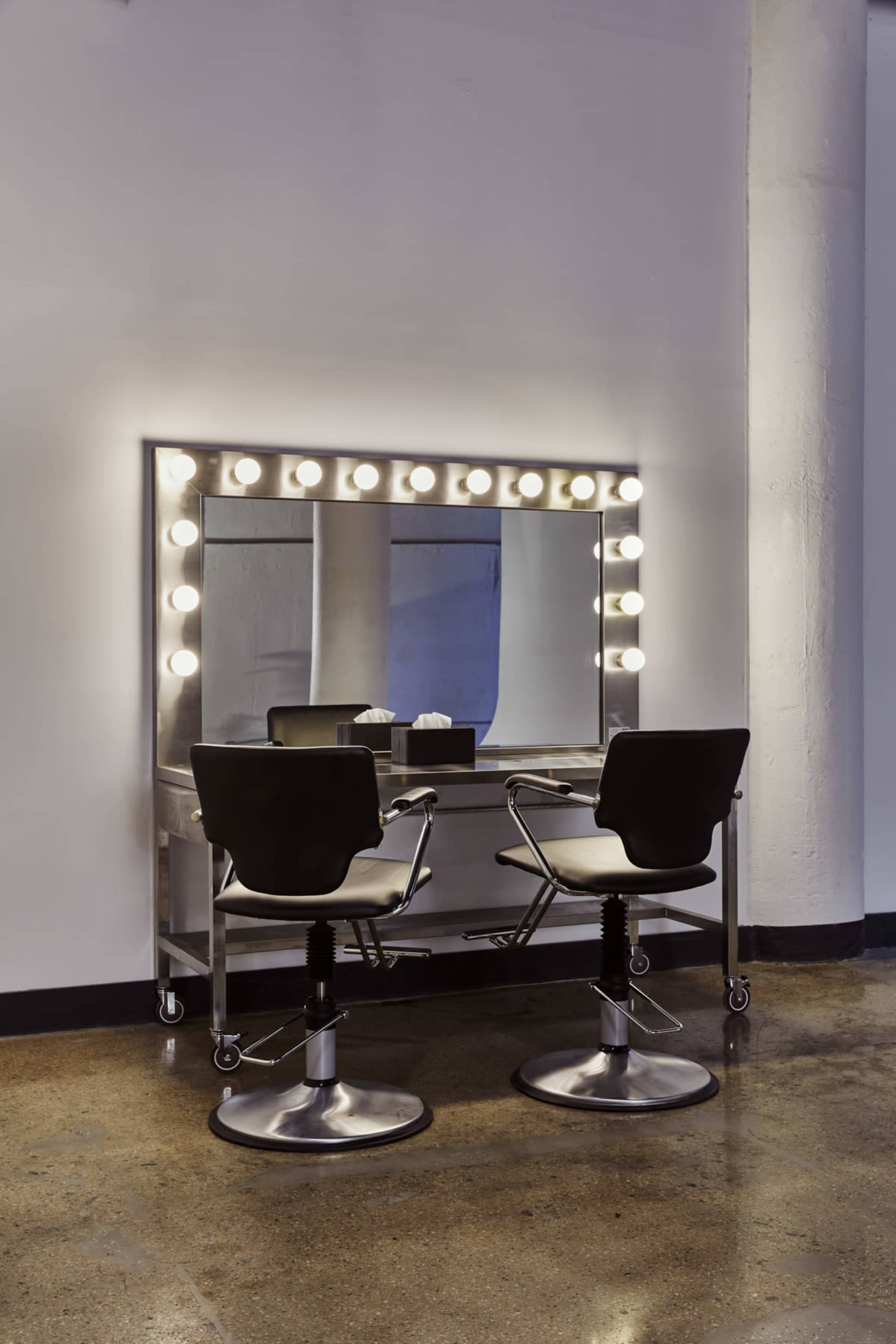 A well-lit makeup vanity with a large mirror and two rolling black chairs stands against a white wall.