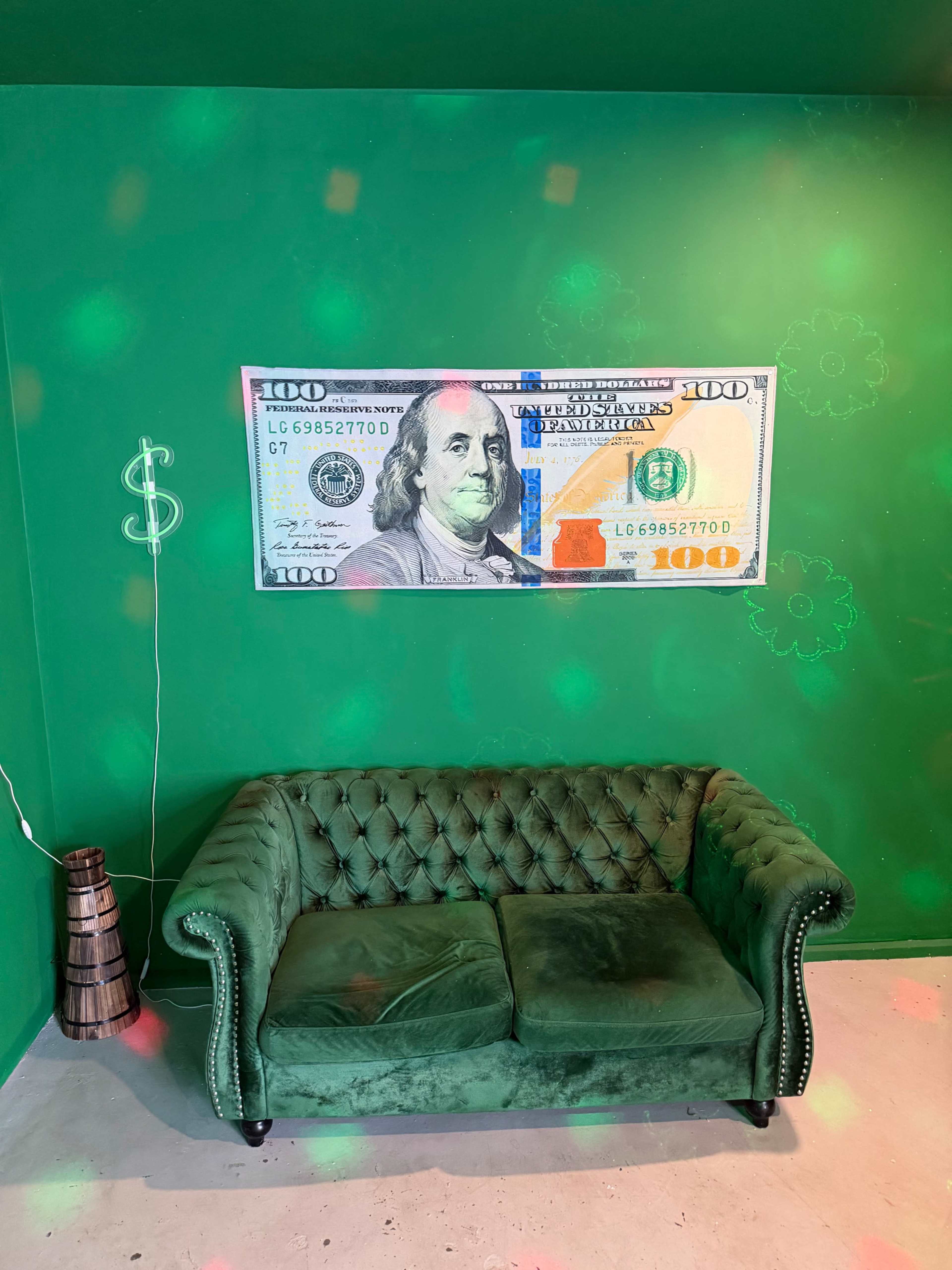 The image features a green room with a green velvet couch and a large framed picture of a $100 bill hanging on the wall.