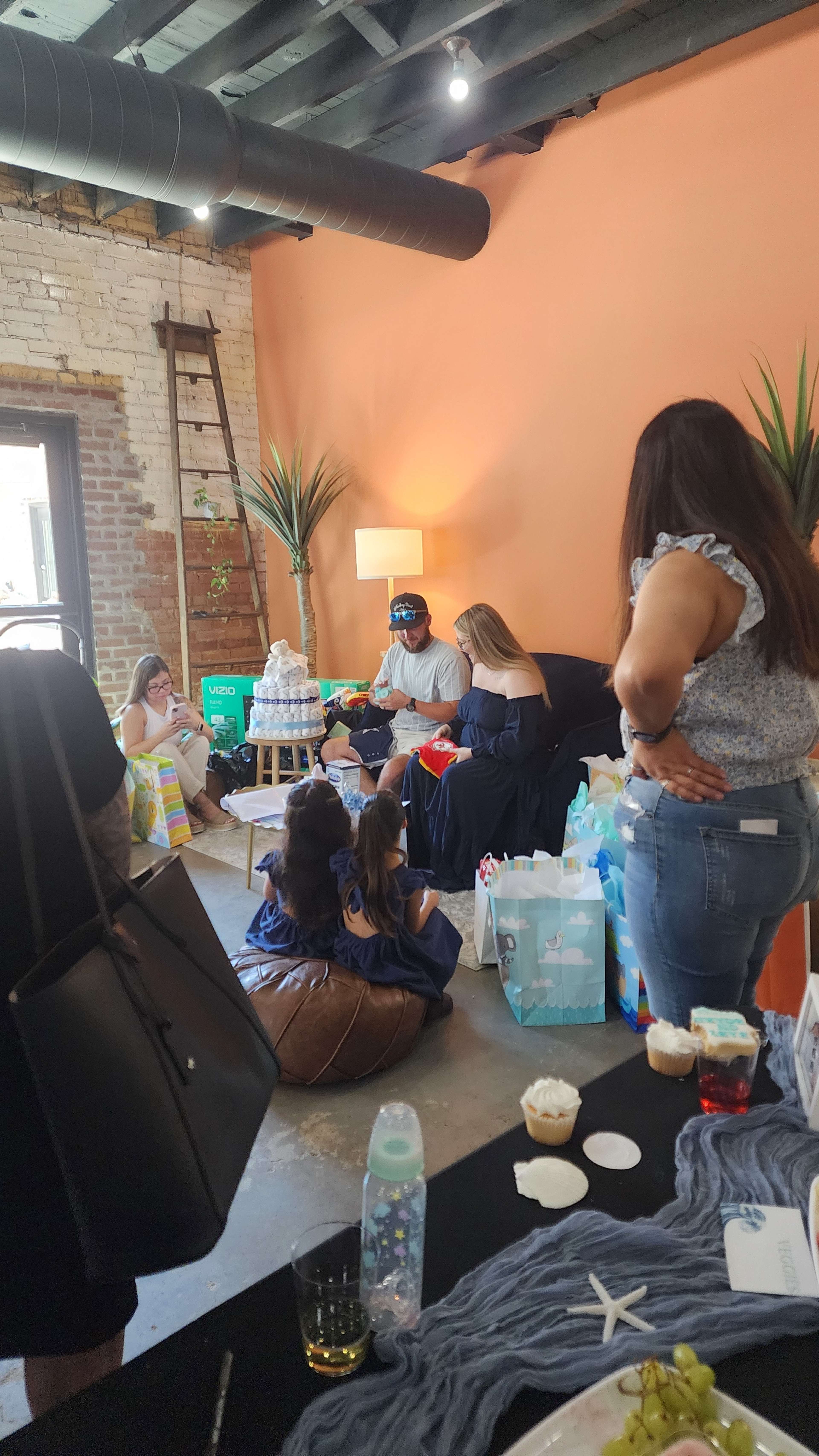 A group of people is gathered in a brightly lit room, sitting on the floor and furniture, surrounded by gifts and refreshments.