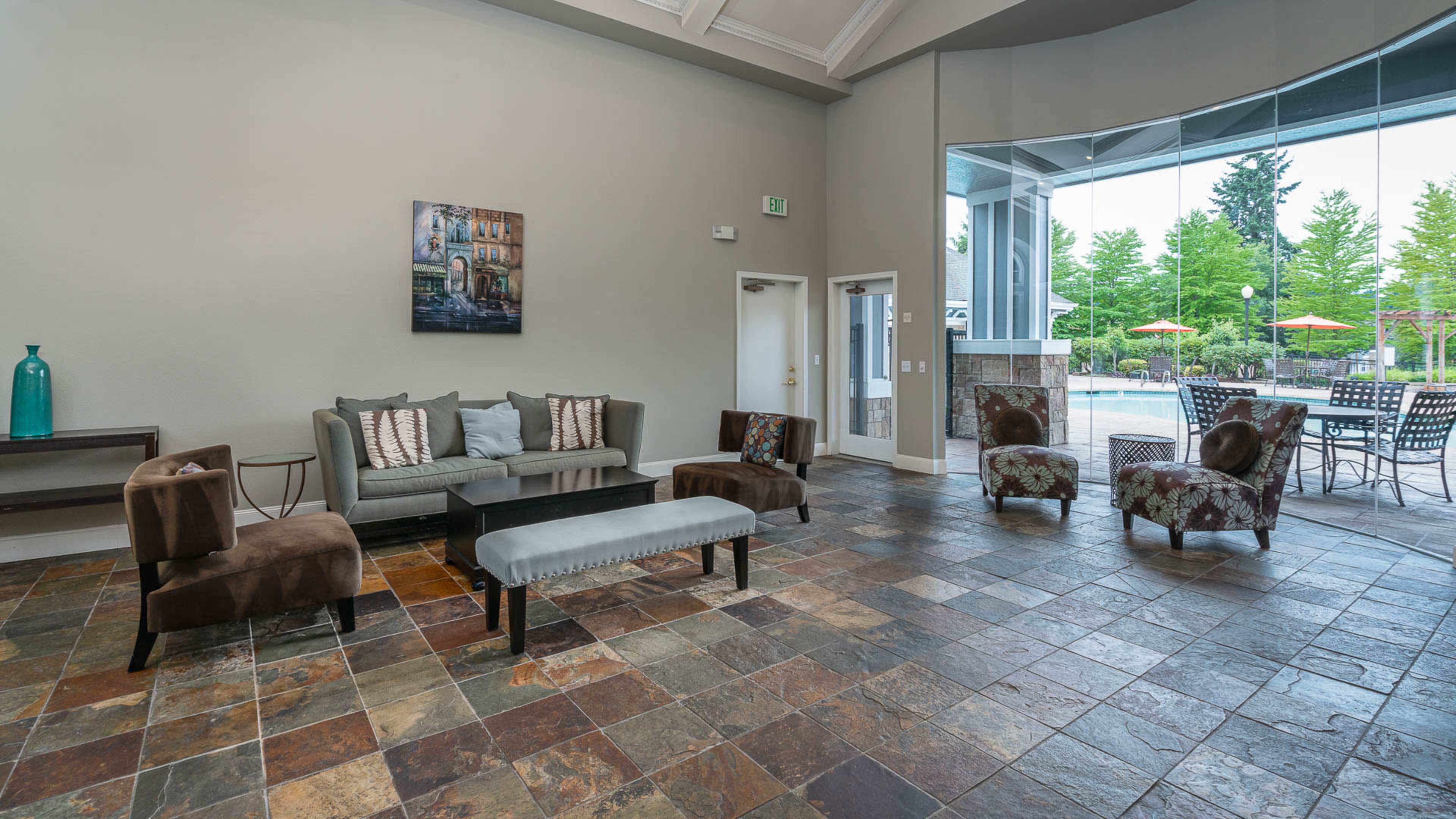 The image shows a spacious lobby area with slate flooring, a gray sofa, upholstered chairs, and large windows overlooking an outdoor patio with tables and umbrellas.