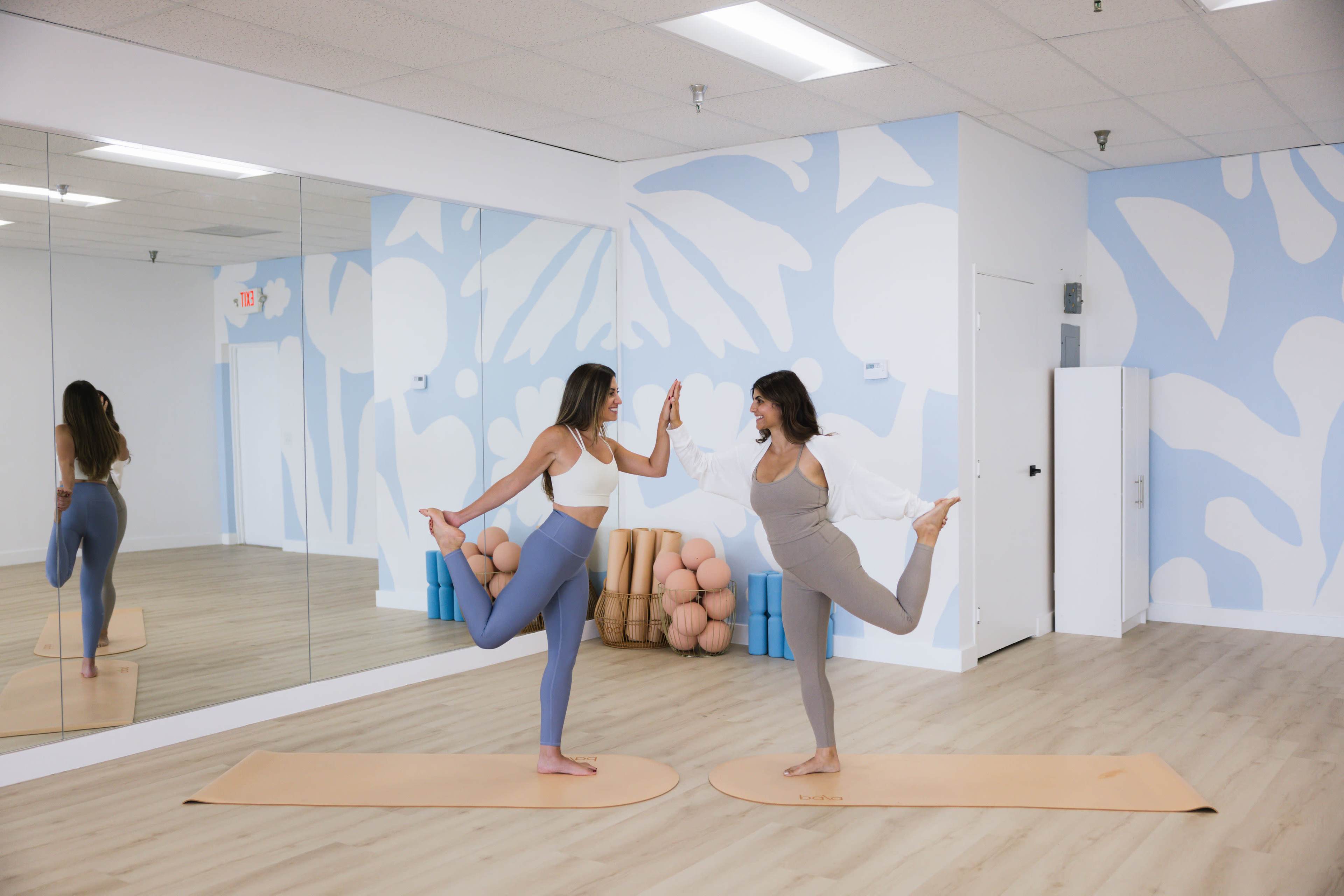 Two women in workout attire perform a yoga pose on mats in a brightly colored exercise studio with a mirrored wall.