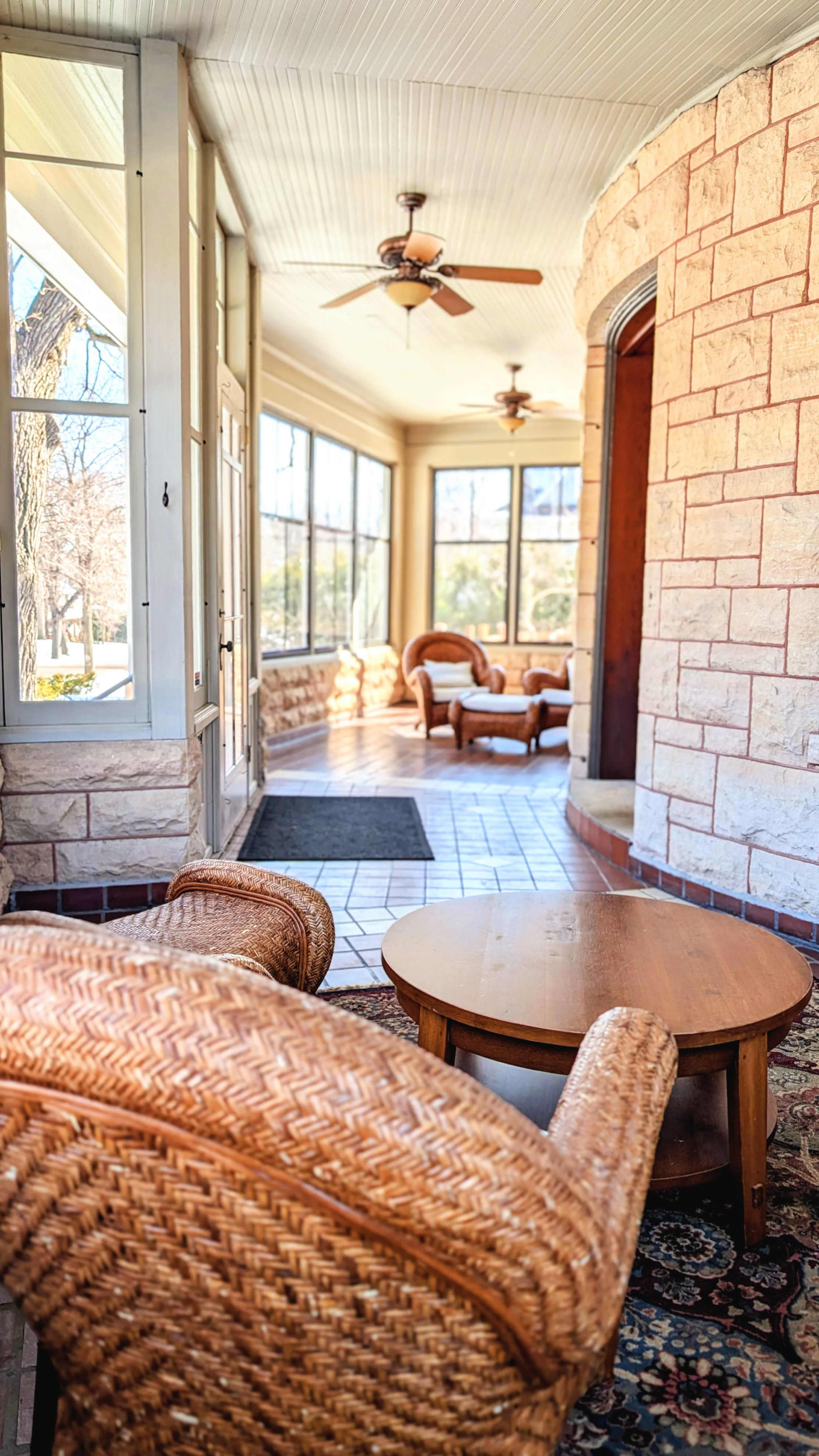 The image shows a sunlit sunroom with stone walls, large windows, and two wicker chairs surrounding a wooden table.