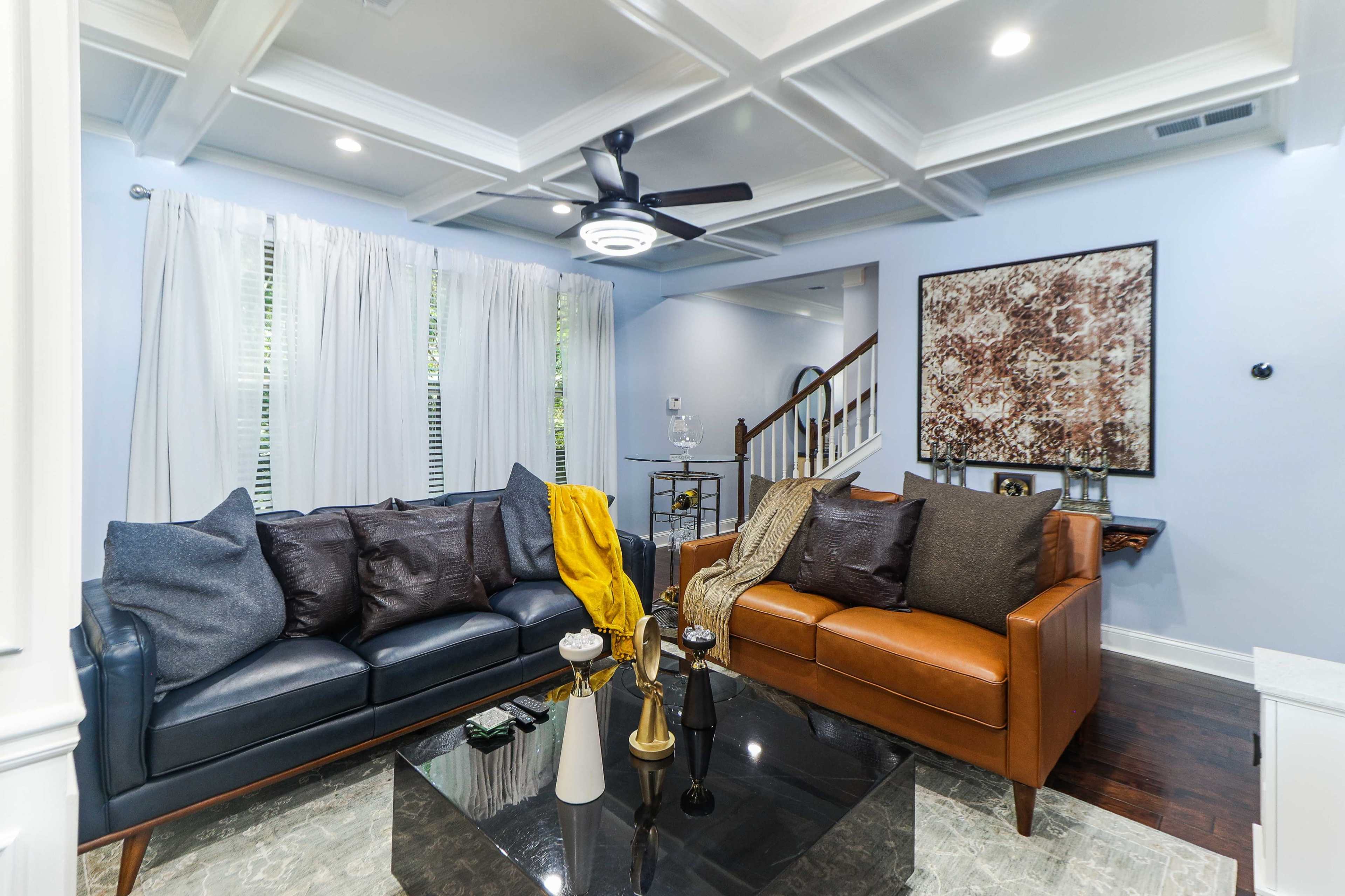 A living room with two sofas, one black and one brown, facing a coffee table on a marble floor, complemented by a decorative wall art and a staircase in the background.