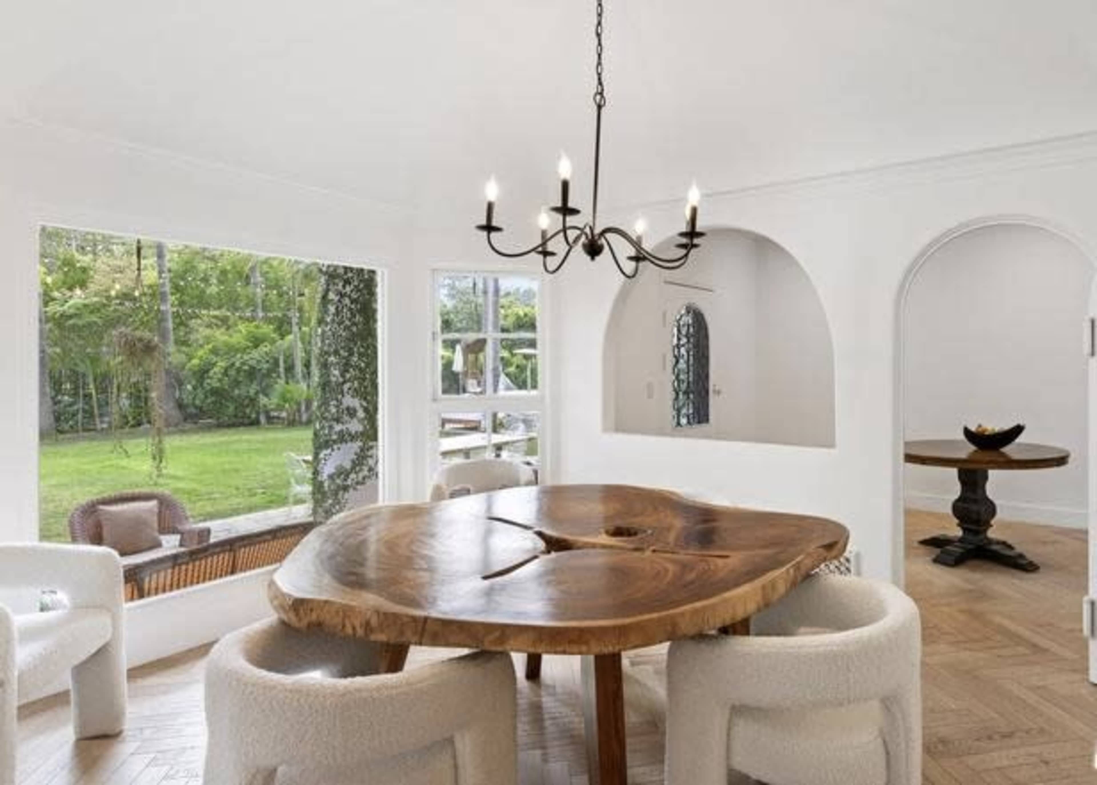The dining area features a large wooden table surrounded by upholstered chairs, with an elegant chandelier overhead and large windows that overlook a green outdoor space.