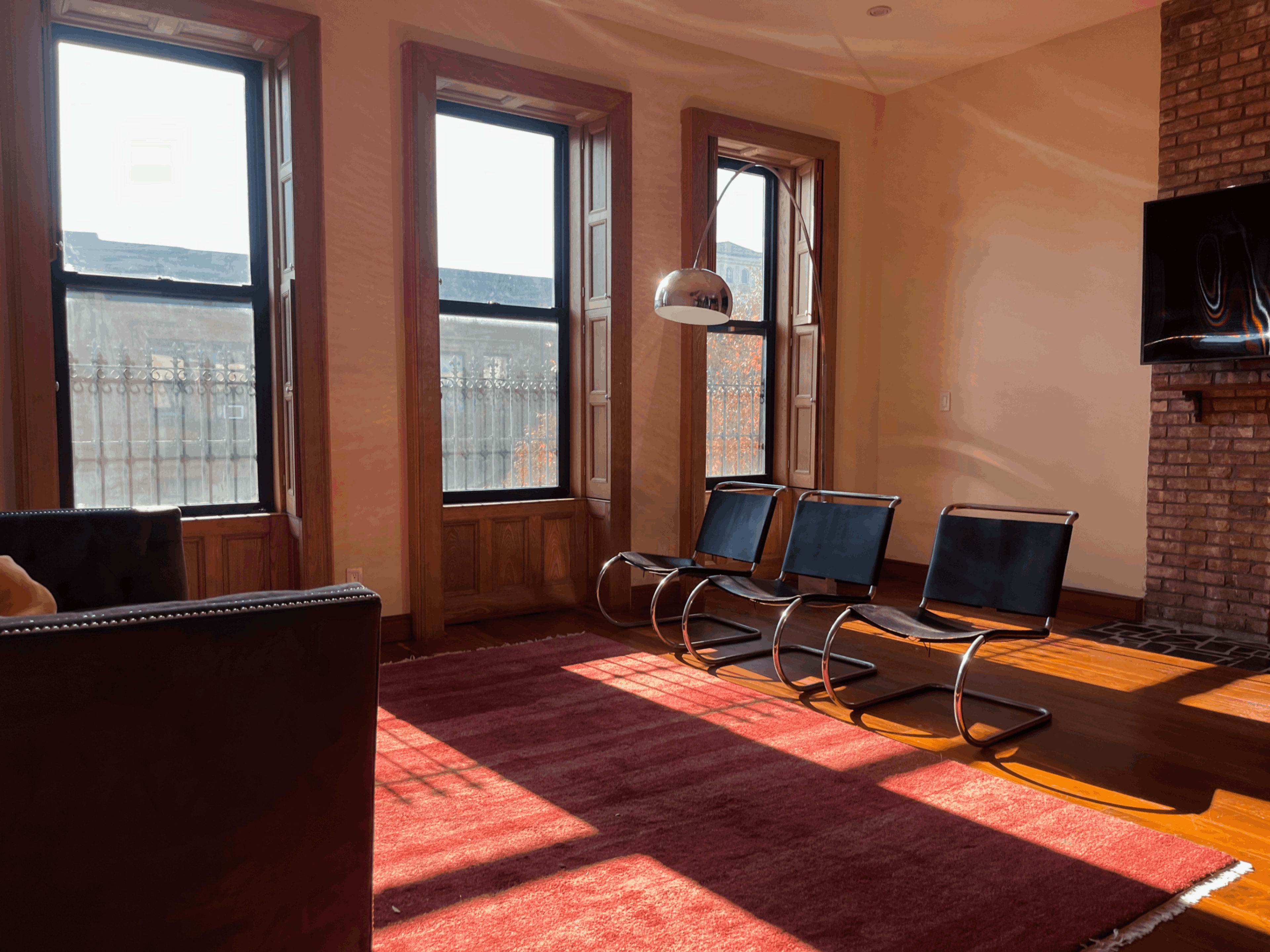 The room features three black chairs positioned on a red rug under sunlight filtering through large windows.