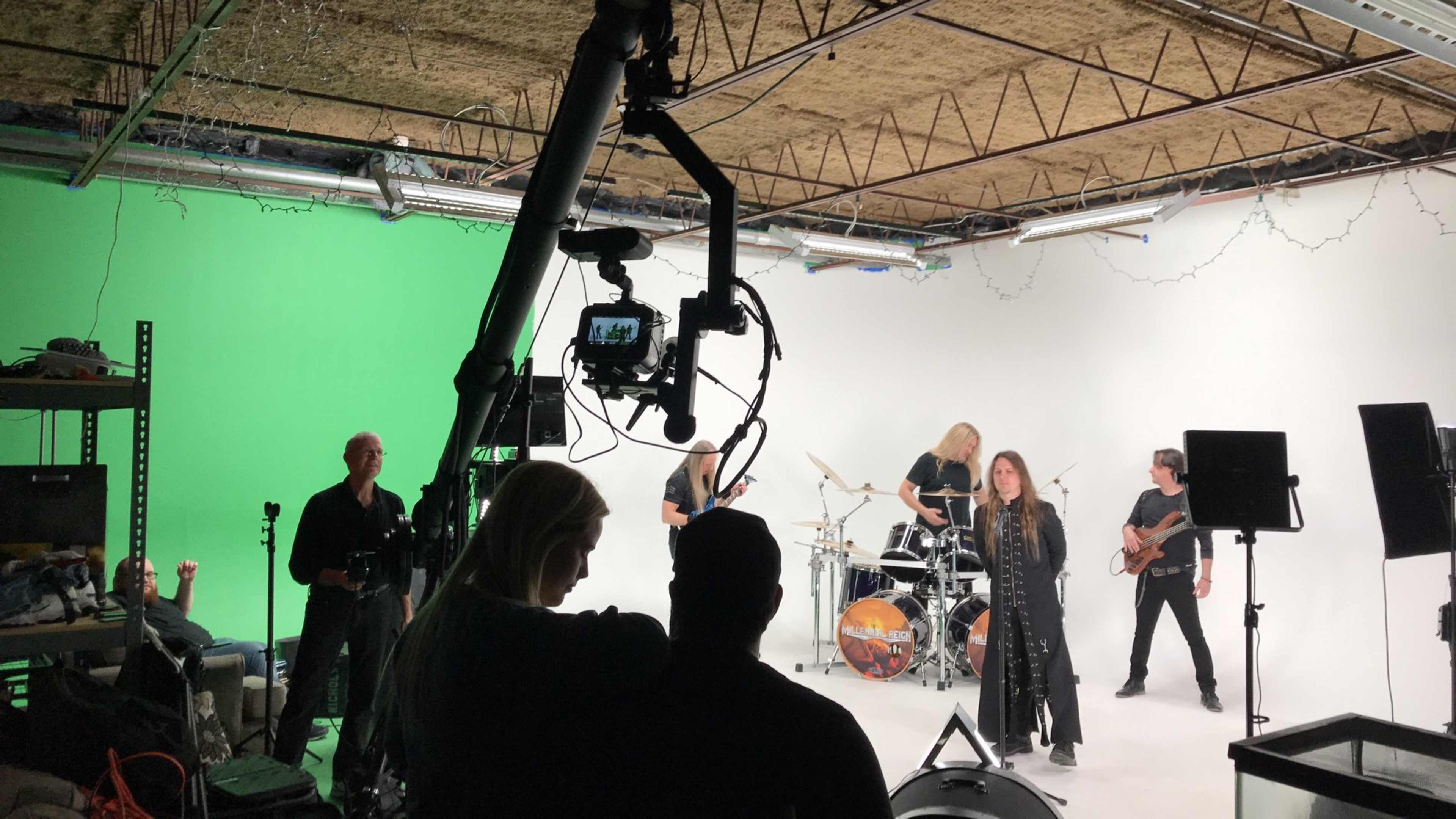 A band performs in a studio with a green screen backdrop while crew members operate cameras and lighting equipment.