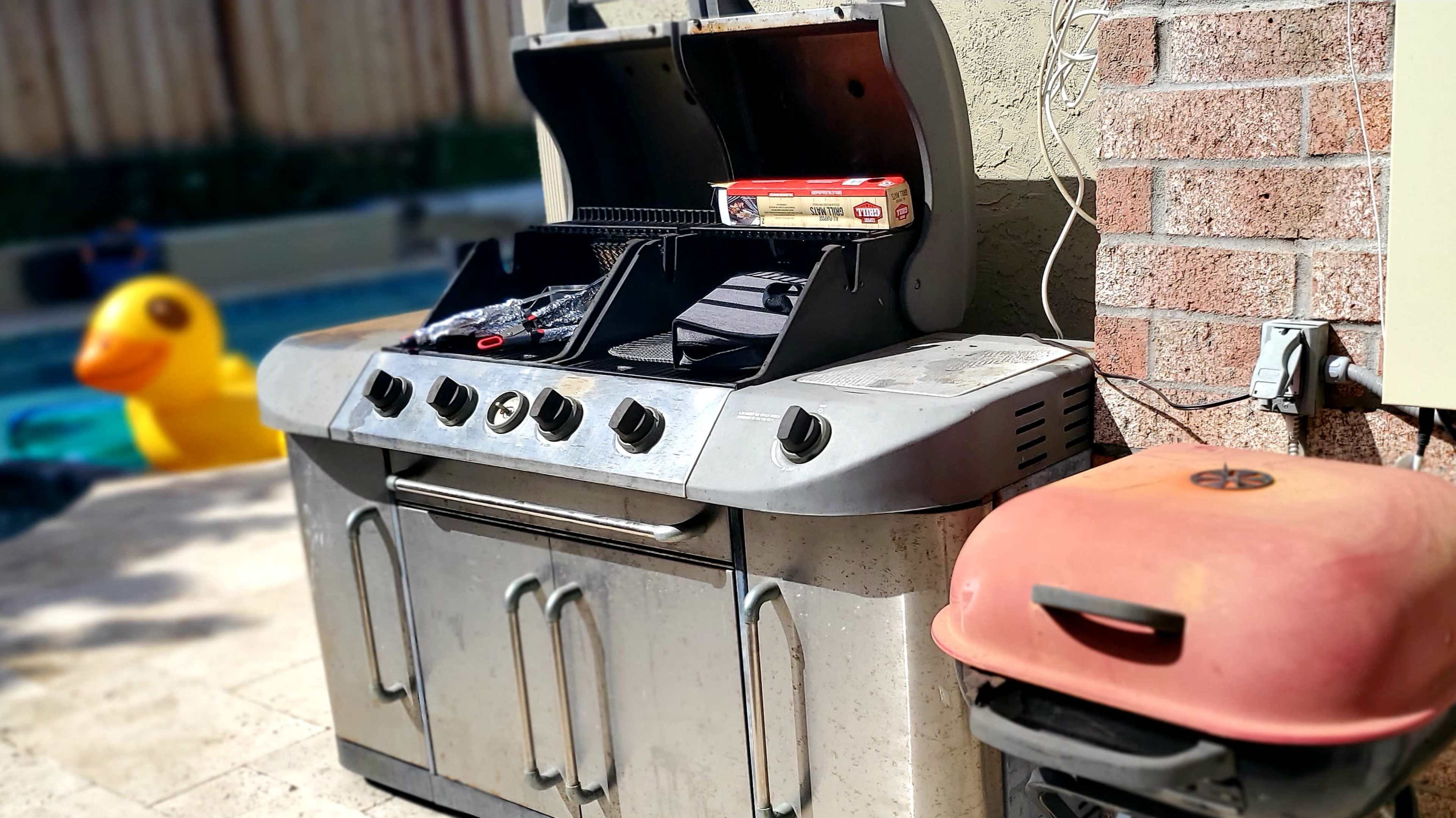 A stainless steel barbecue grill next to a red gas grill, with a large rubber duck visible in the background near a pool.