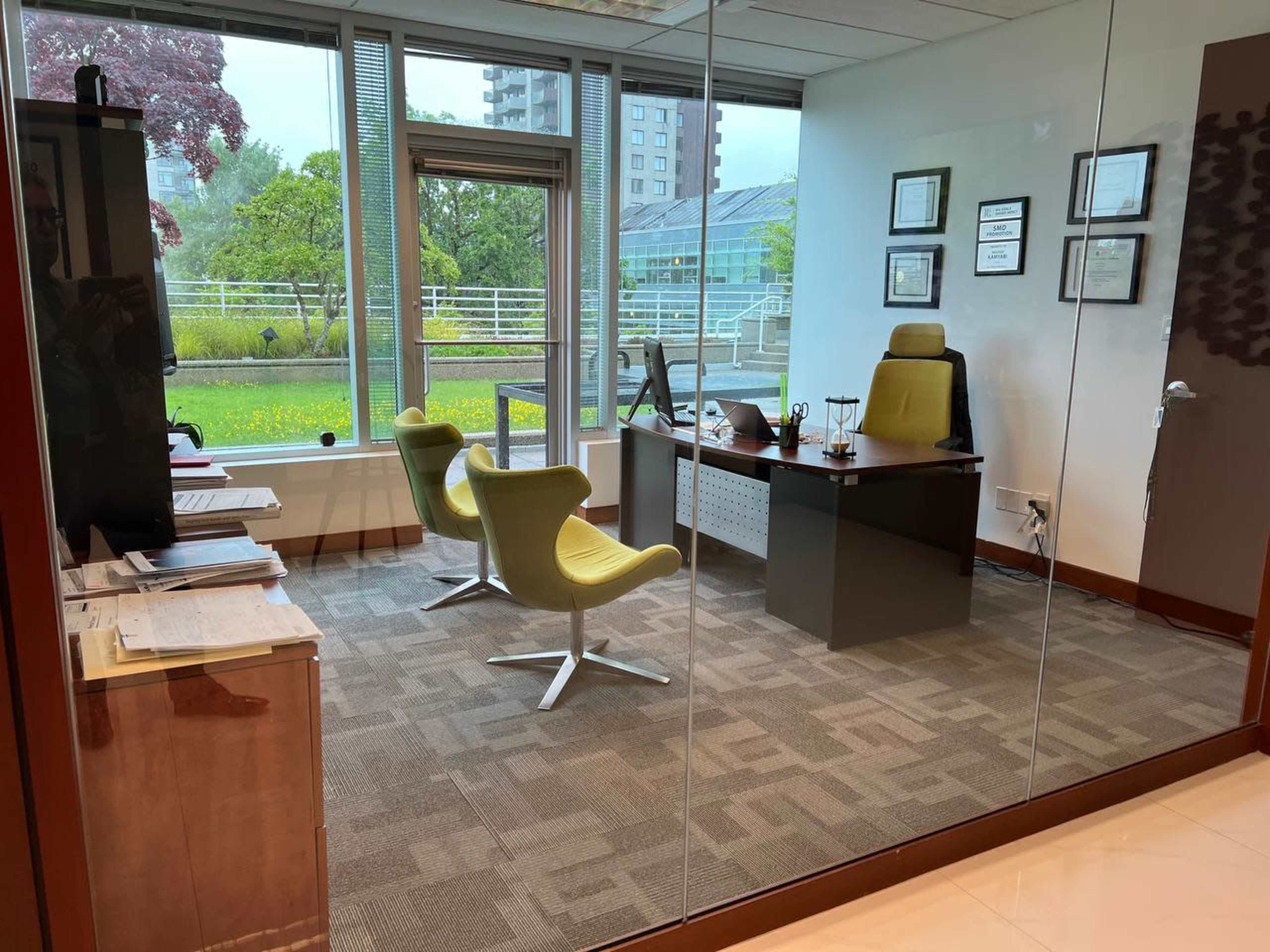 A modern office space with a glass wall, featuring a desk, two chairs, framed documents, and a view of greenery outside.