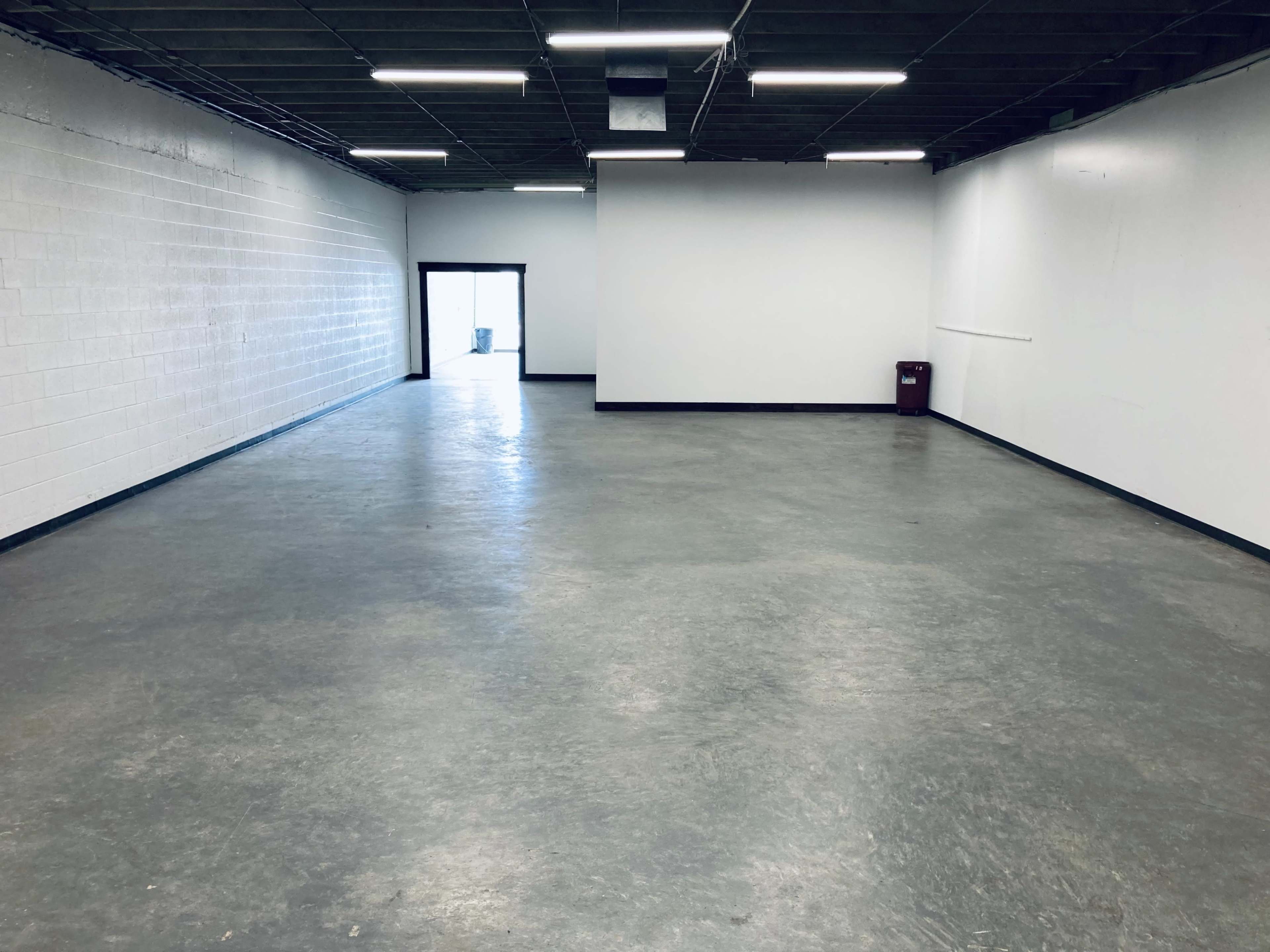 An empty room with concrete flooring and white walls, featuring a door at the far end and a trash can in one corner.