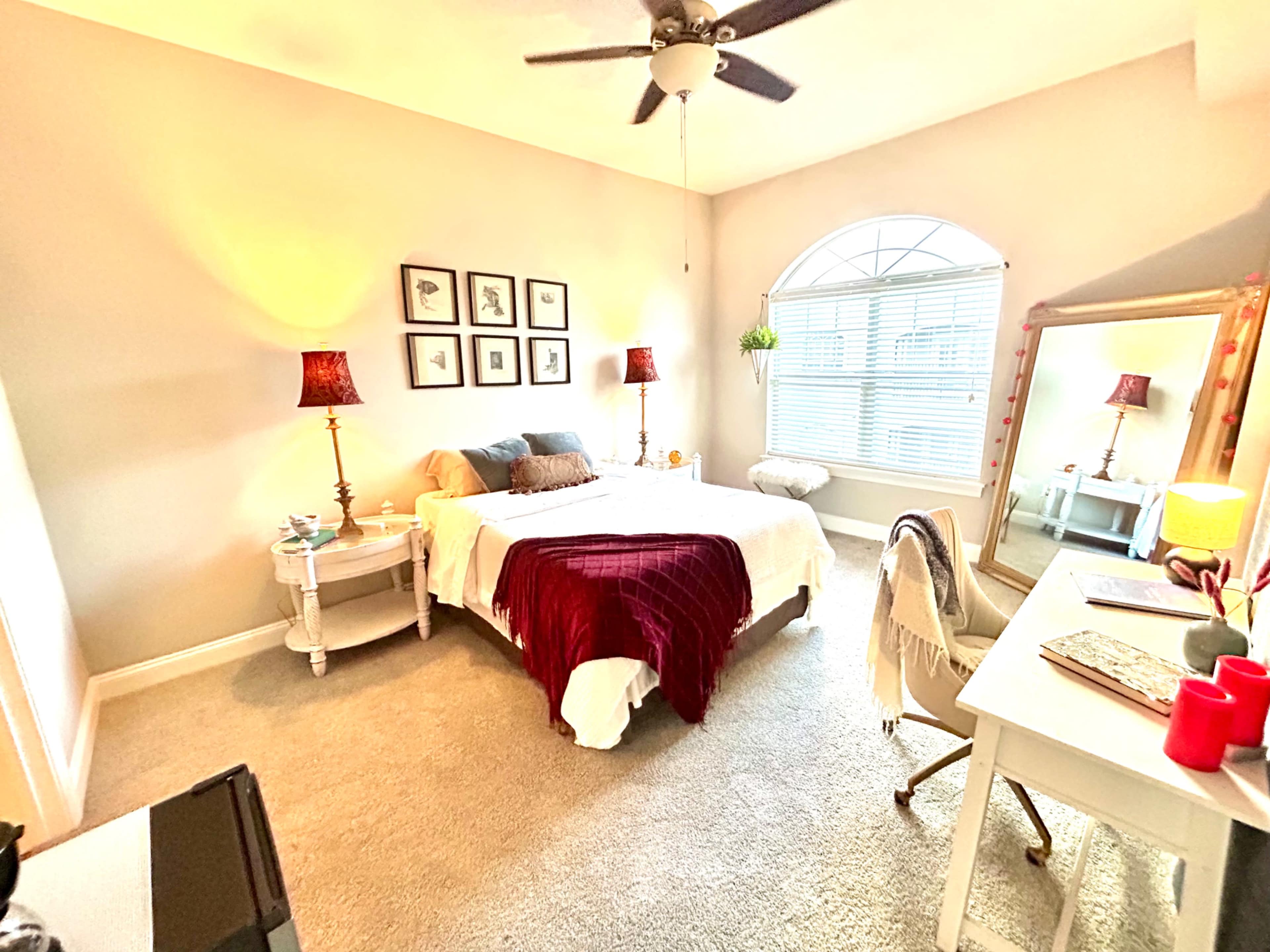 The image shows a well-lit bedroom featuring a queen bed with a maroon throw, two bedside lamps, a large mirror, and a desk with a chair.