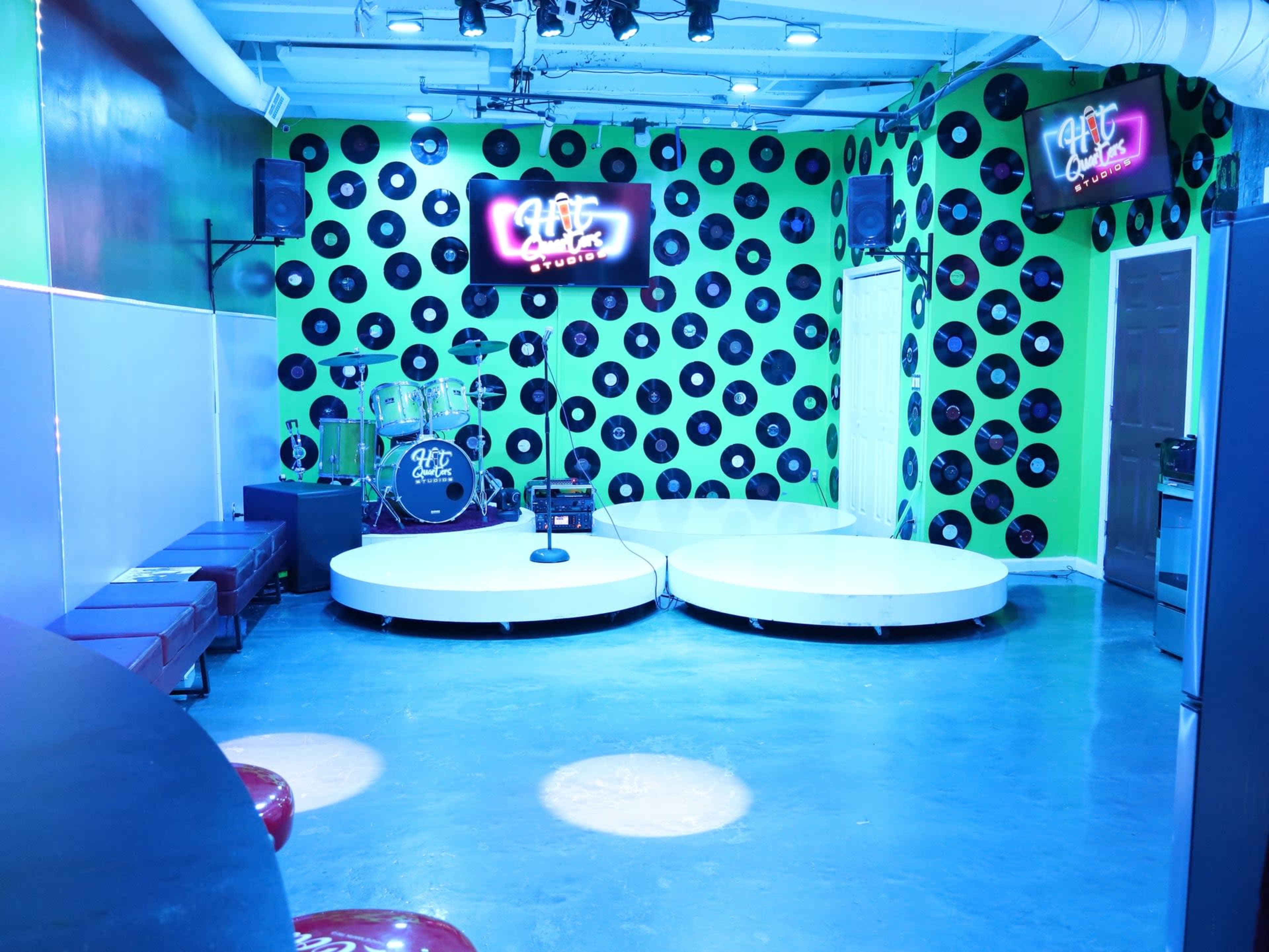 The image depicts a music venue with a vibrant green and black decor featuring vinyl records on the walls, a drum set on stage, and two circular platforms for performances.