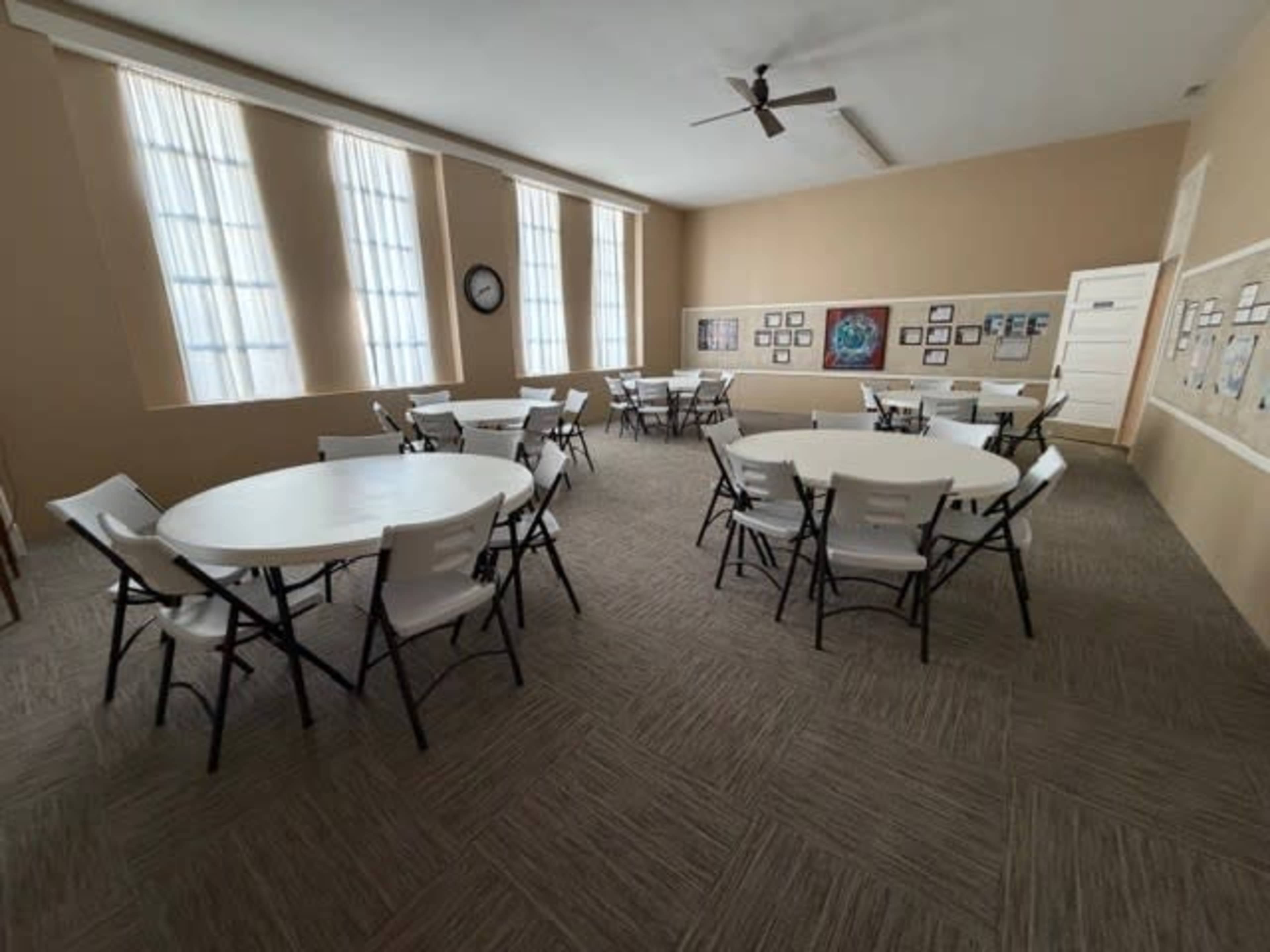 The image shows a large room with several round tables surrounded by folding chairs, illuminated by light coming through tall windows.